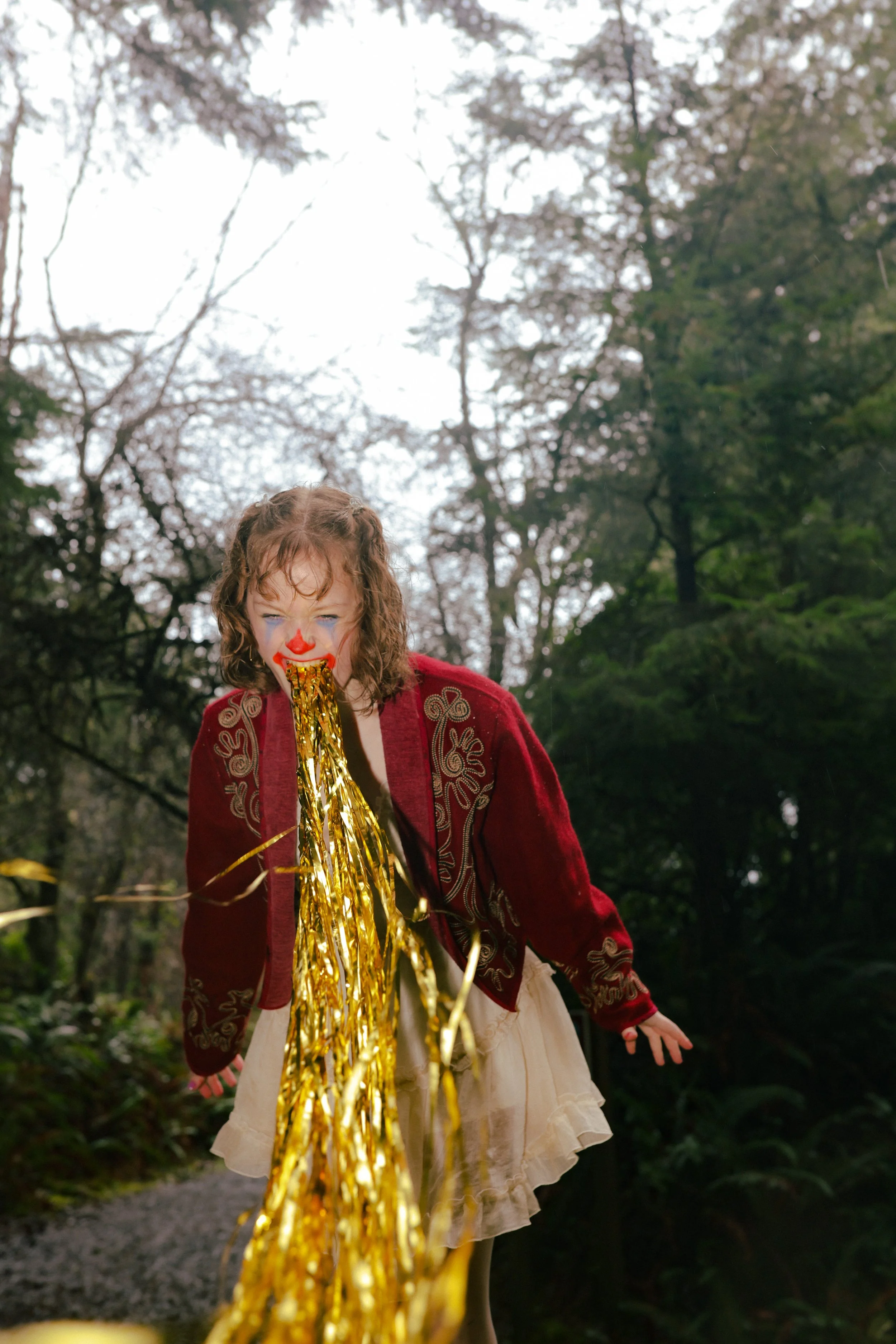 A young girl with curly hair and clown makeup on her face, dressed in a red jacket and a beige dress, sticking her tongue out with gold tinsel around it in a forested area.