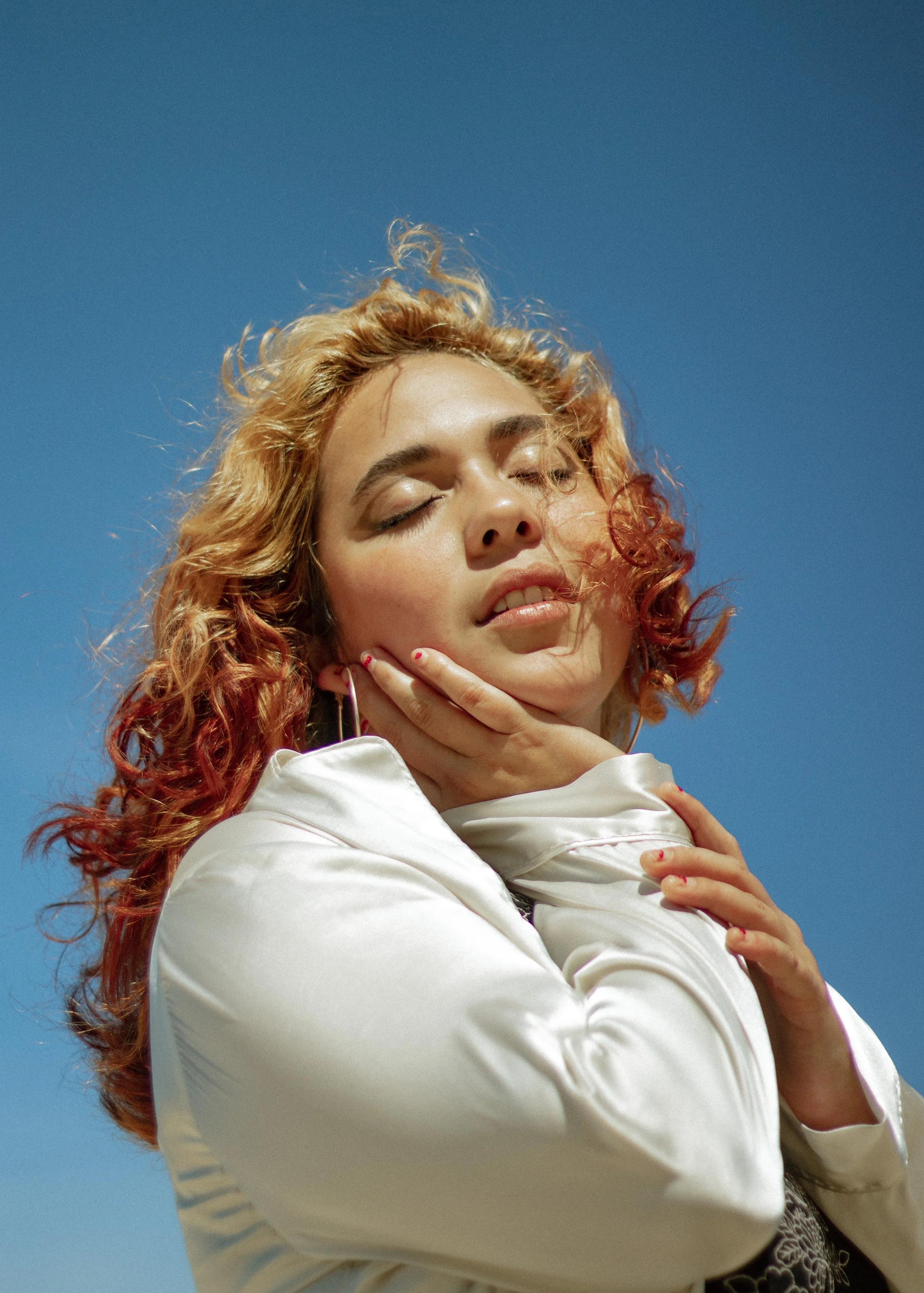 A young woman with curly red hair, closed eyes, and a serene expression, posing against a bright blue sky with her hands gently touching her face and shoulder.
