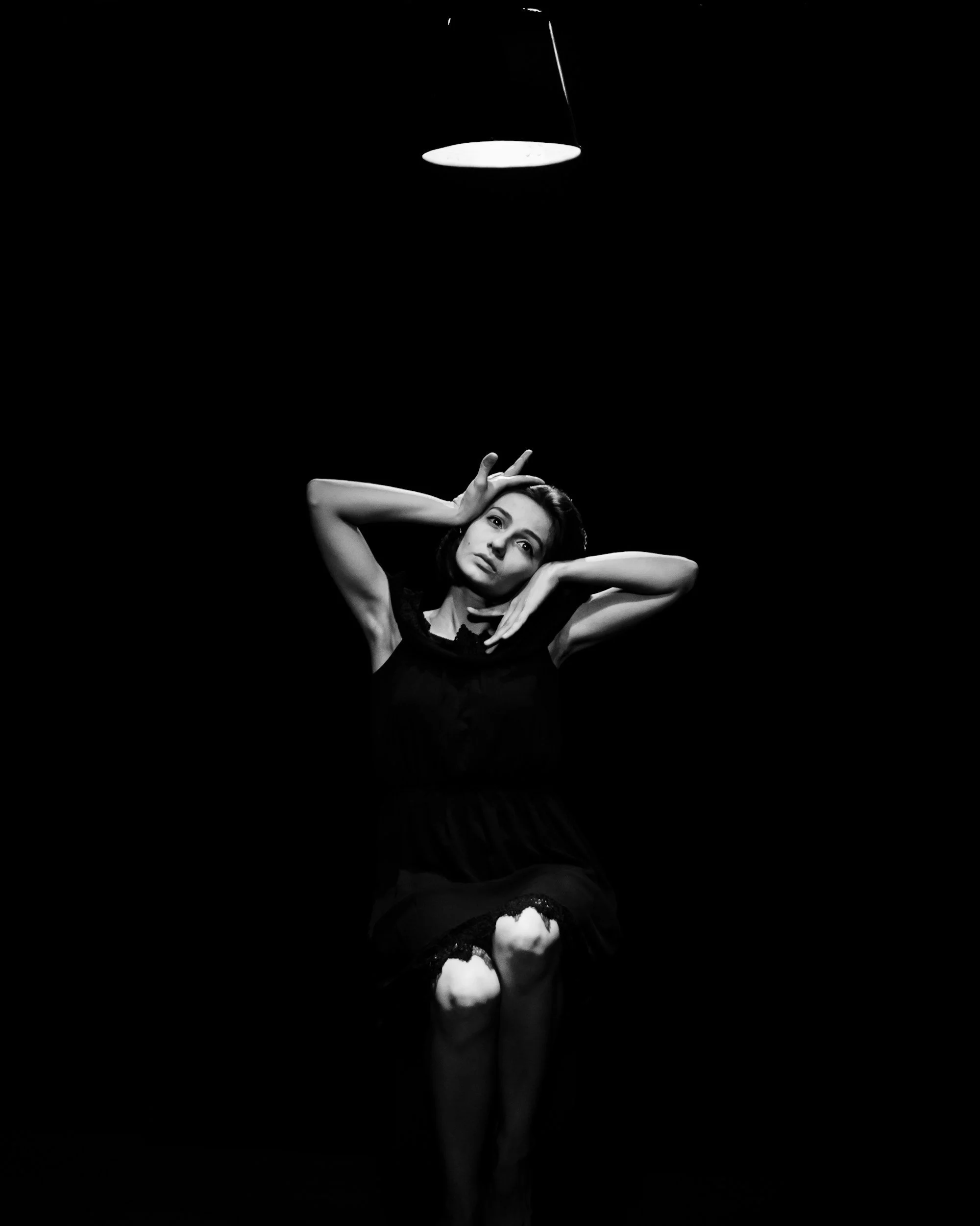 A woman sitting with her knees pulled up, posing under a hanging light in a dark room. The image is in black and white.