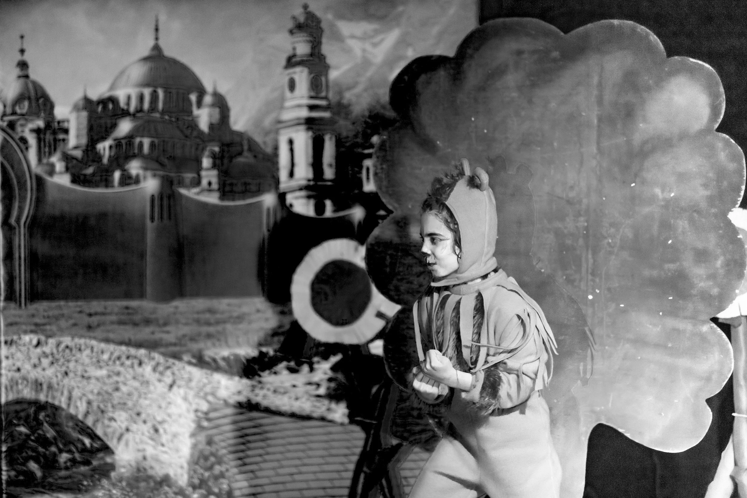 A young woman wearing a hoodie with ears, standing in front of a painted backdrop of a castle and a cloud of smoke, holding an object in her hand.