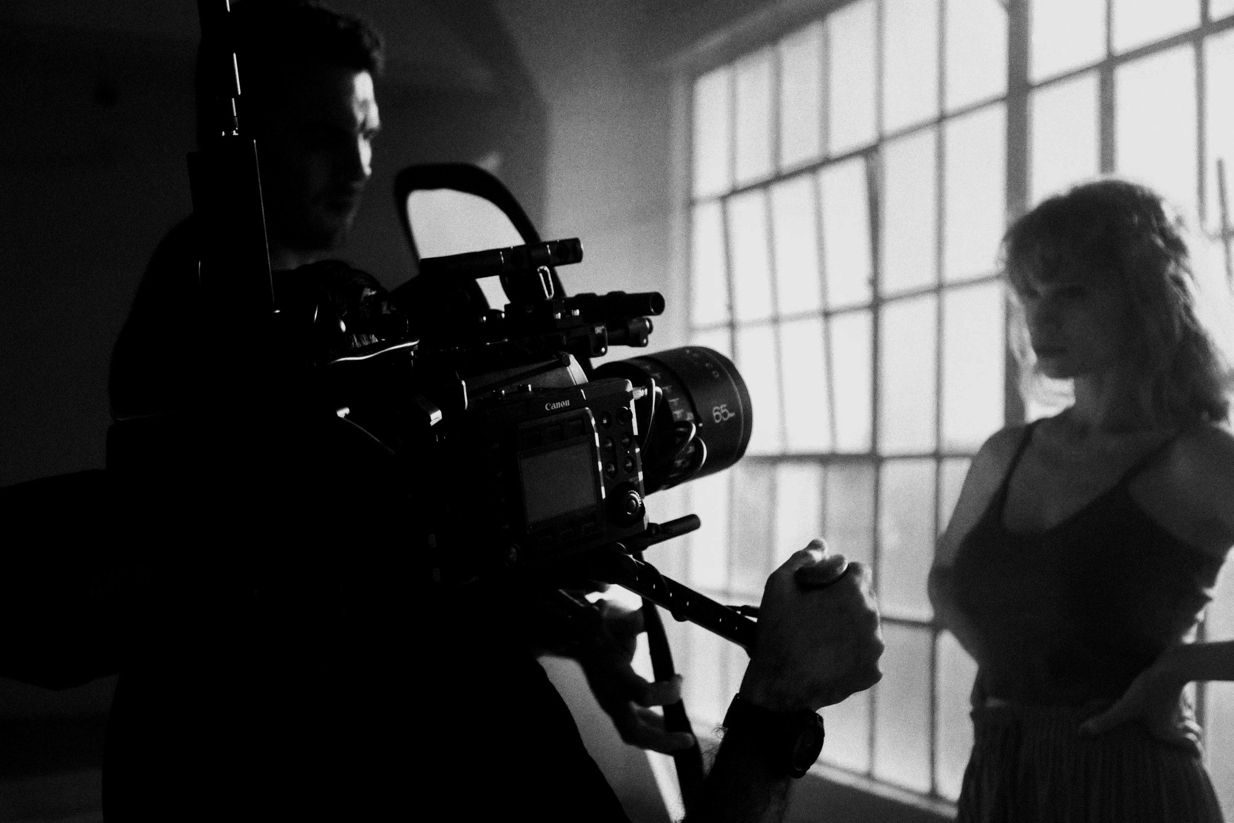 A person in a striped shirt sits on a stool during a photoshoot, with a photographer using a camera on a tripod capturing the scene, all in black and white.