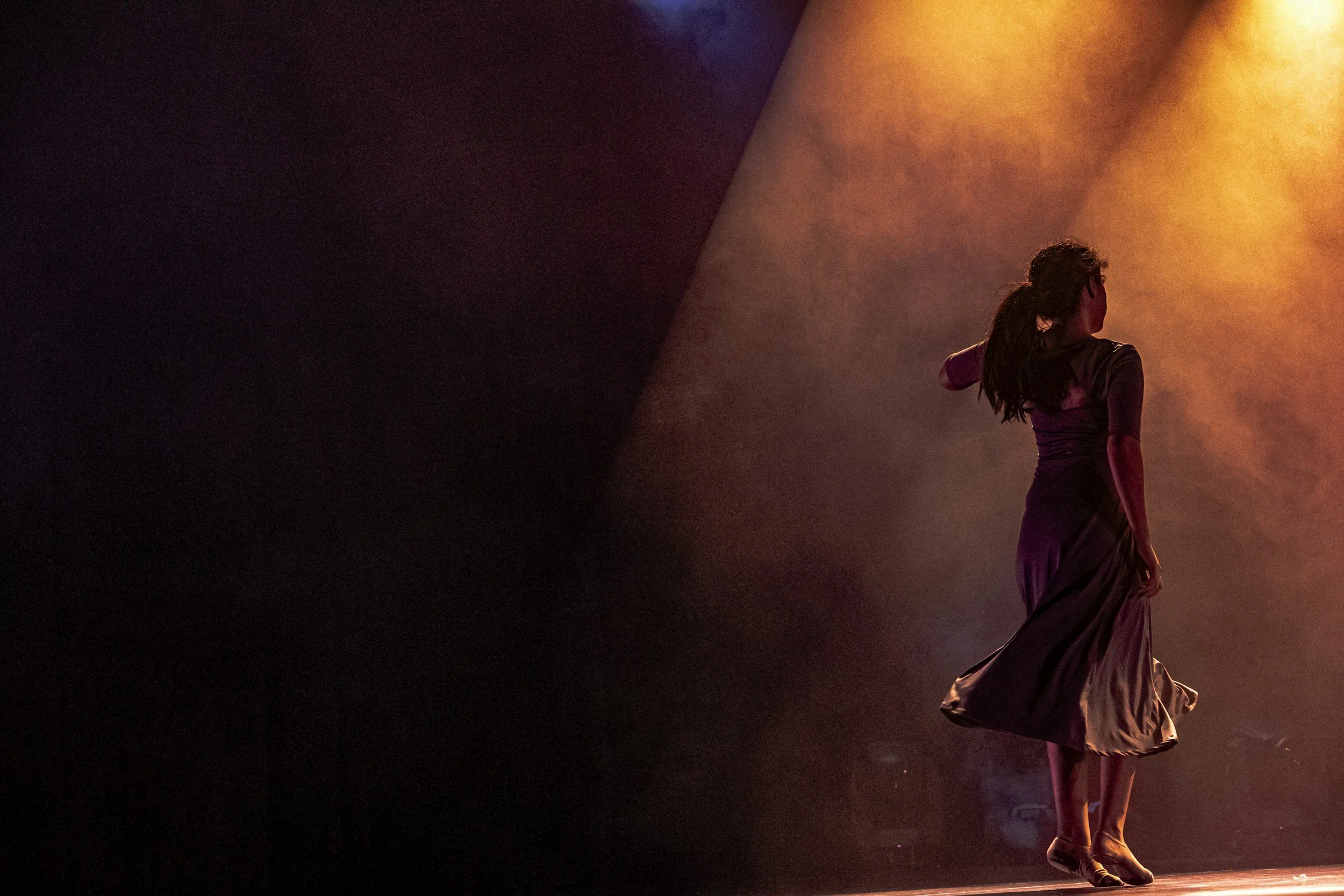 A woman with long hair wearing a dress, walking on a stage with colored lights and fog, facing away from the camera.