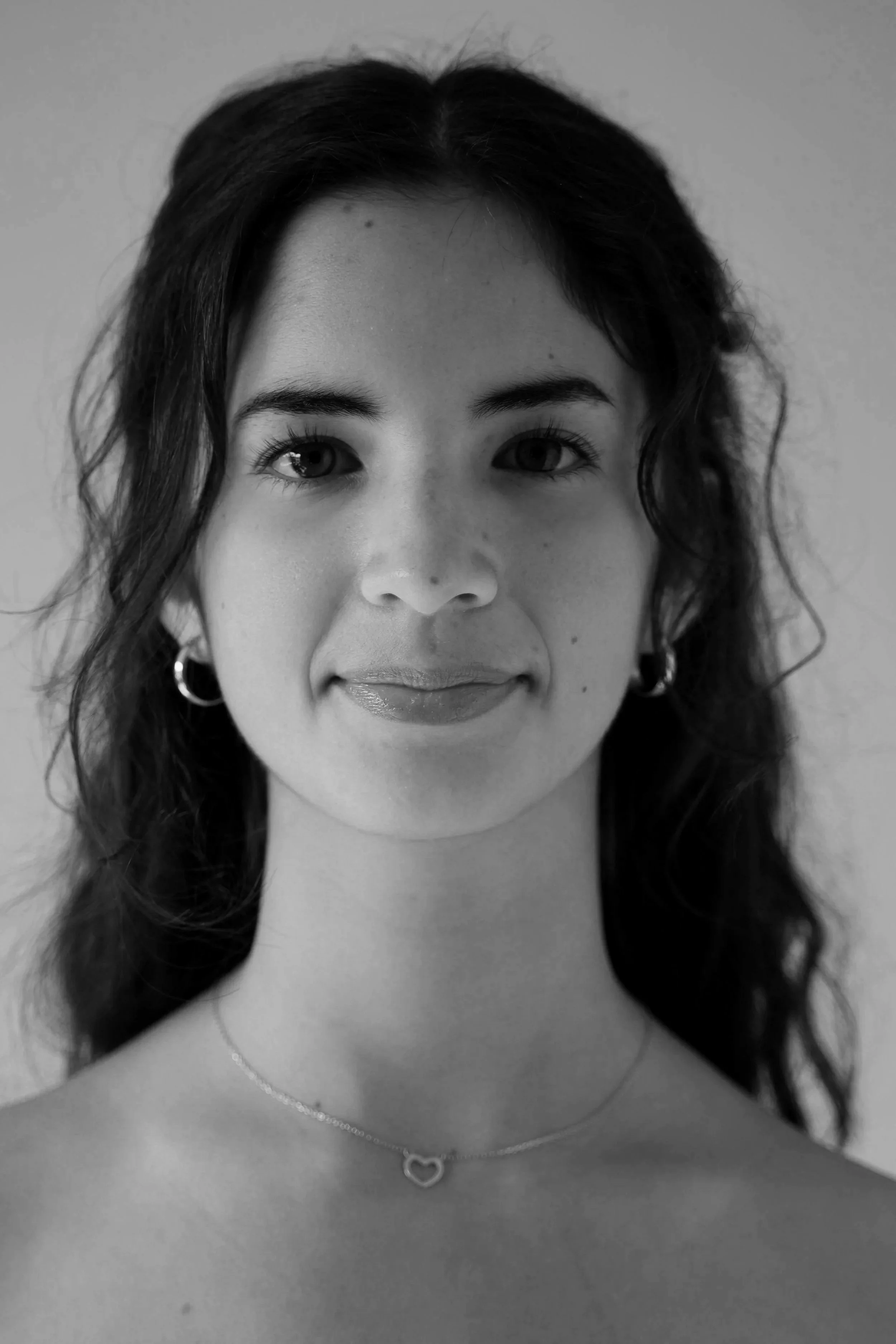 Black-and-white photo of a woman with long wavy hair, wearing hoop earrings and a heart-shaped necklace, smiling slightly