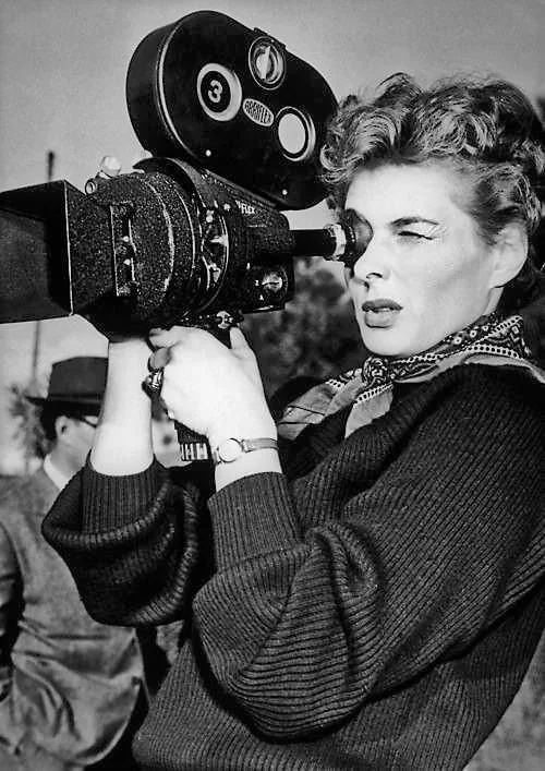 A woman operating a vintage movie camera outdoors, with a man in a hat in the background.
