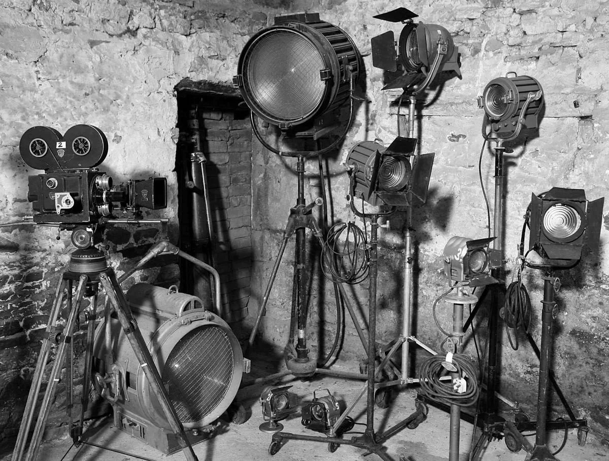 Collection of vintage film lighting equipment including spotlights, lamps, and a film camera, arranged against a brick and stone wall in a studio or storage space.