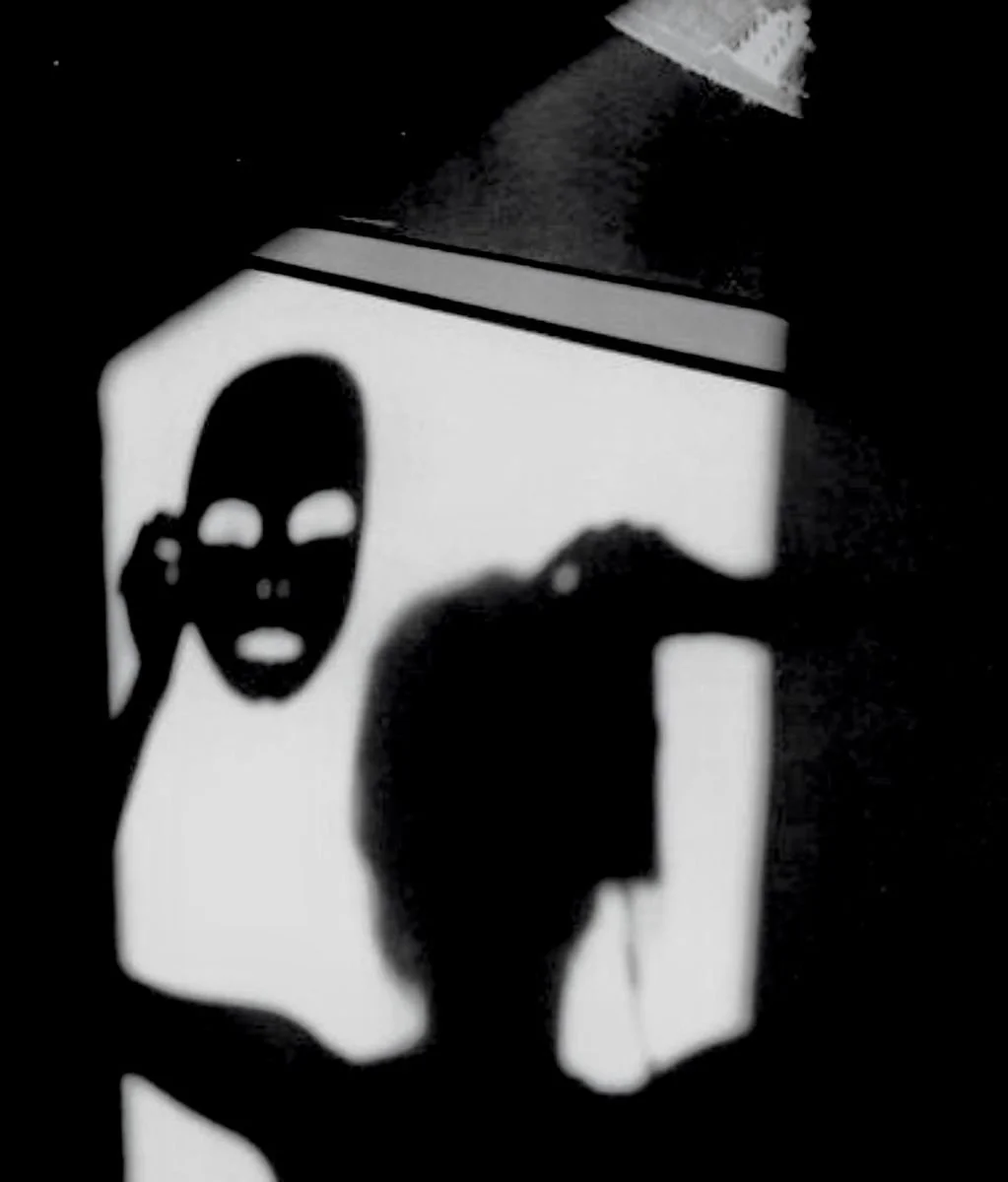 Silhouette of a person taking a mirror selfie with a phone, with a projected image of a mask with hollow eyes and mouth on the wall behind them.