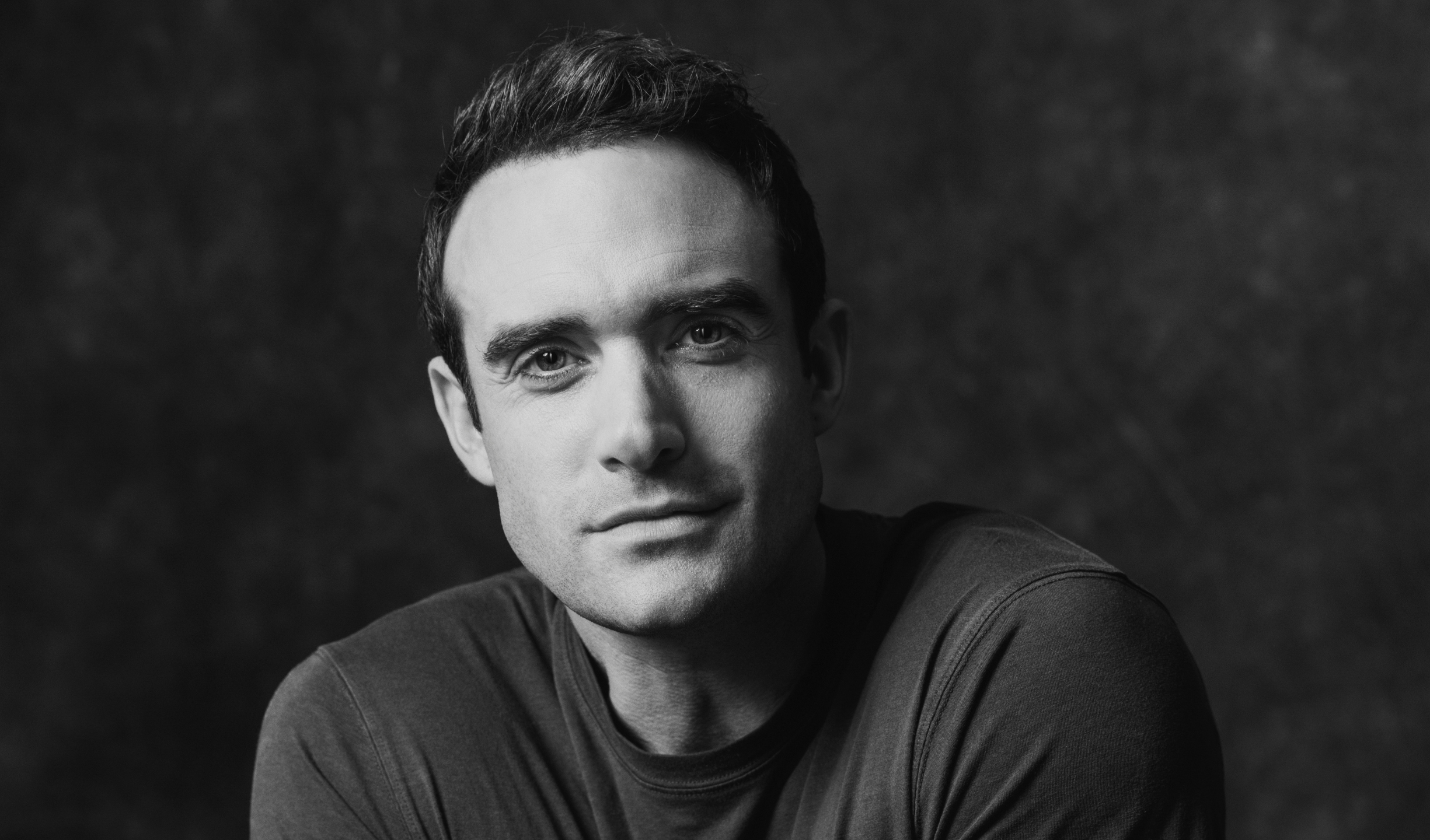 Black and white portrait of a young man with short dark hair, wearing a dark shirt, looking directly at the camera against a dark background.