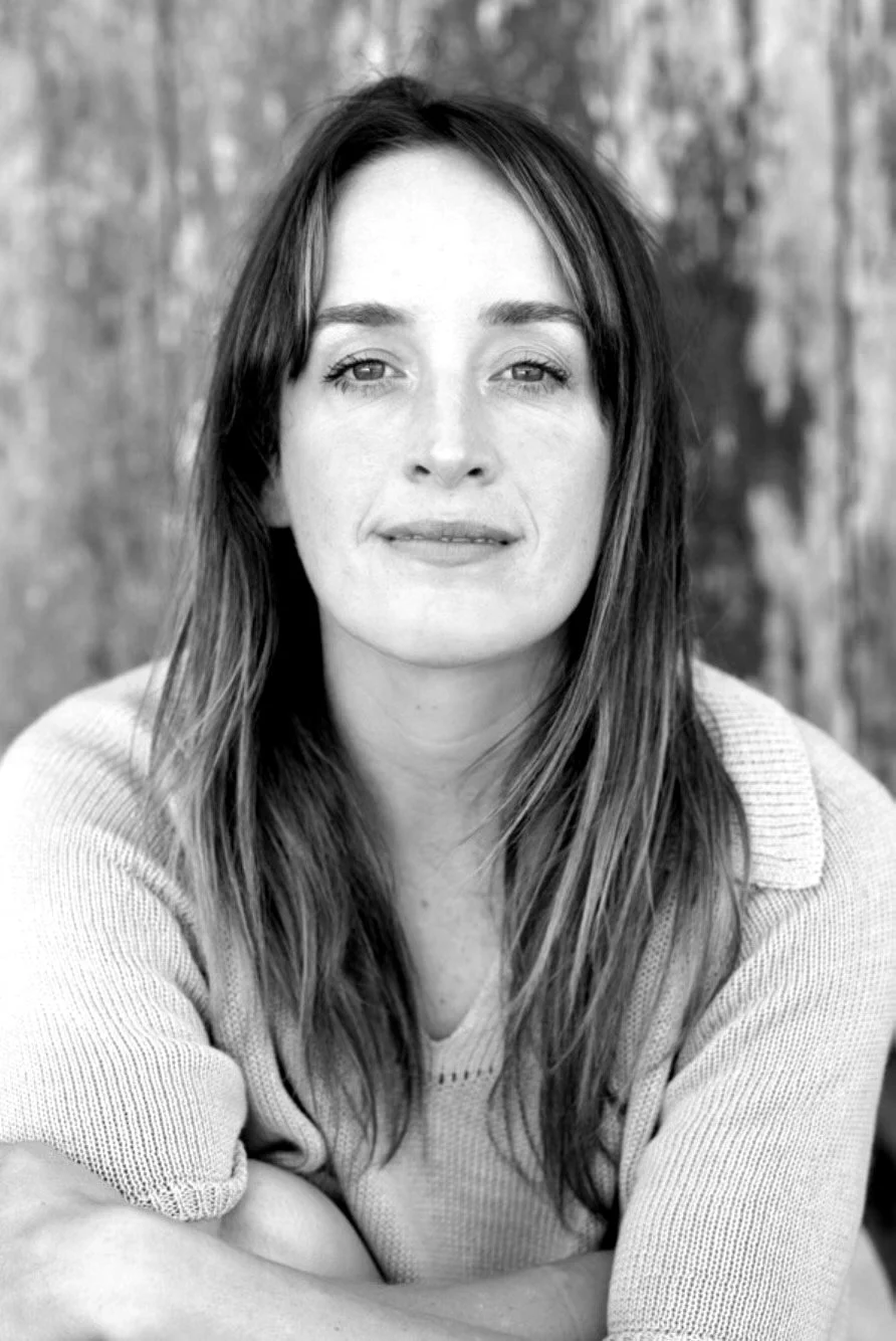 A black and white portrait of a woman with long hair, looking at the camera with a soft expression, outdoors in front of a blurred background.