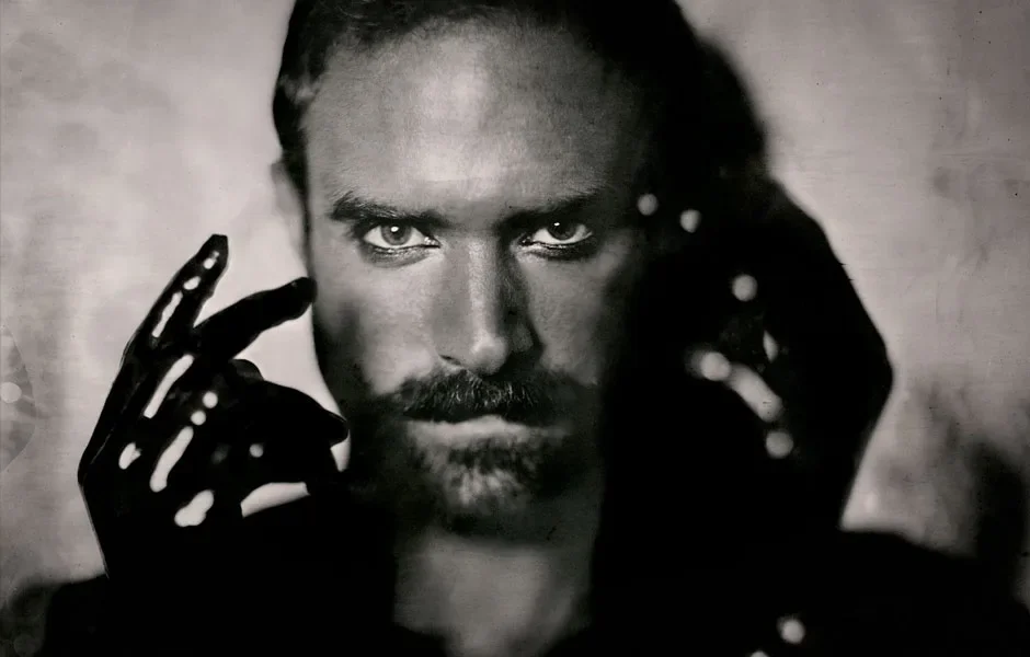 Black and white close-up portrait of a man with intense eyes, mustache, and beard, holding black gloves with white dots near his face.