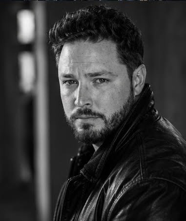 Black and white portrait of a man with short curly hair and a beard, wearing a leather jacket, looking at the camera with a serious expression.