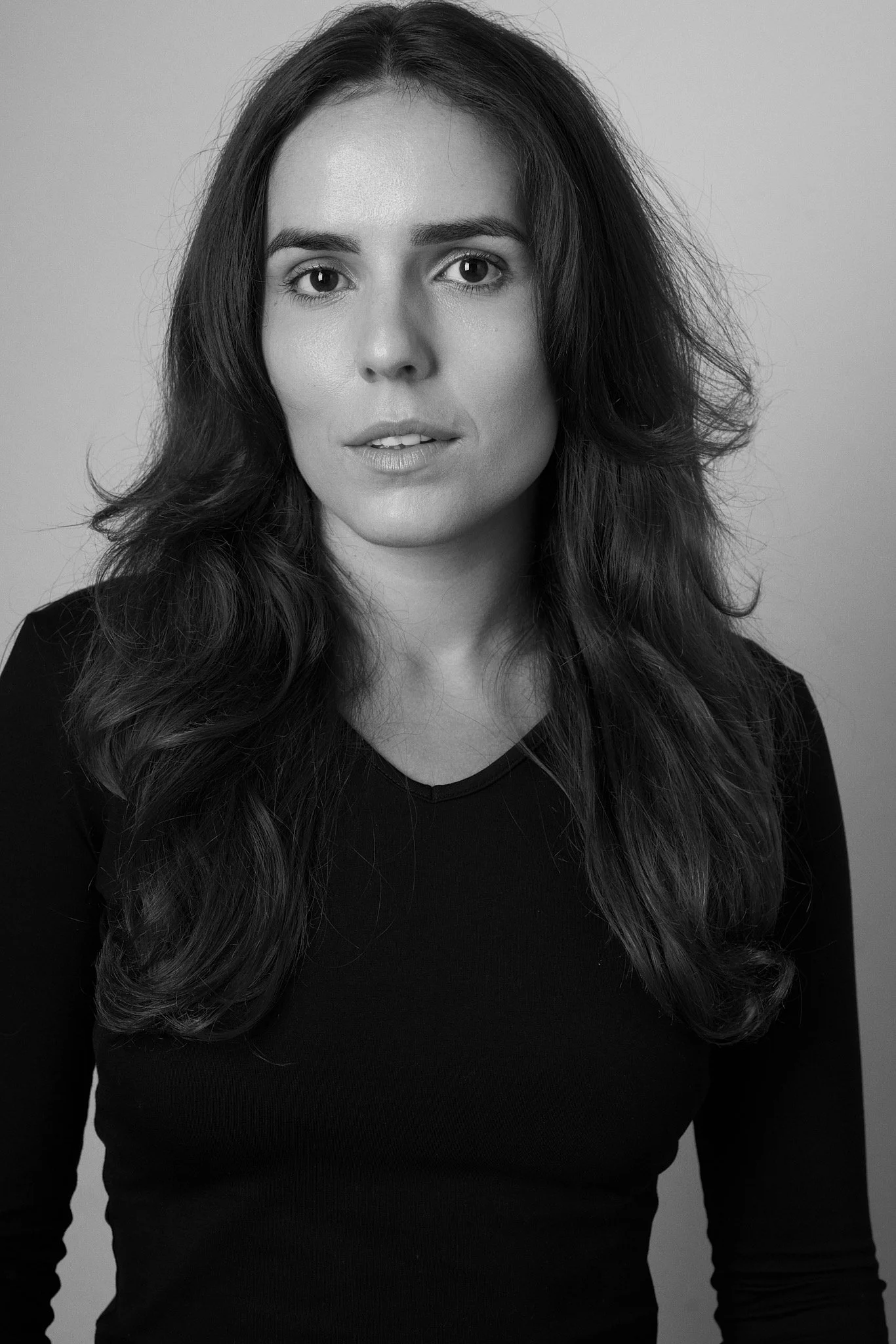 Black and white portrait of a woman with long, wavy hair, wearing a dark top, looking directly at the camera with a neutral expression.