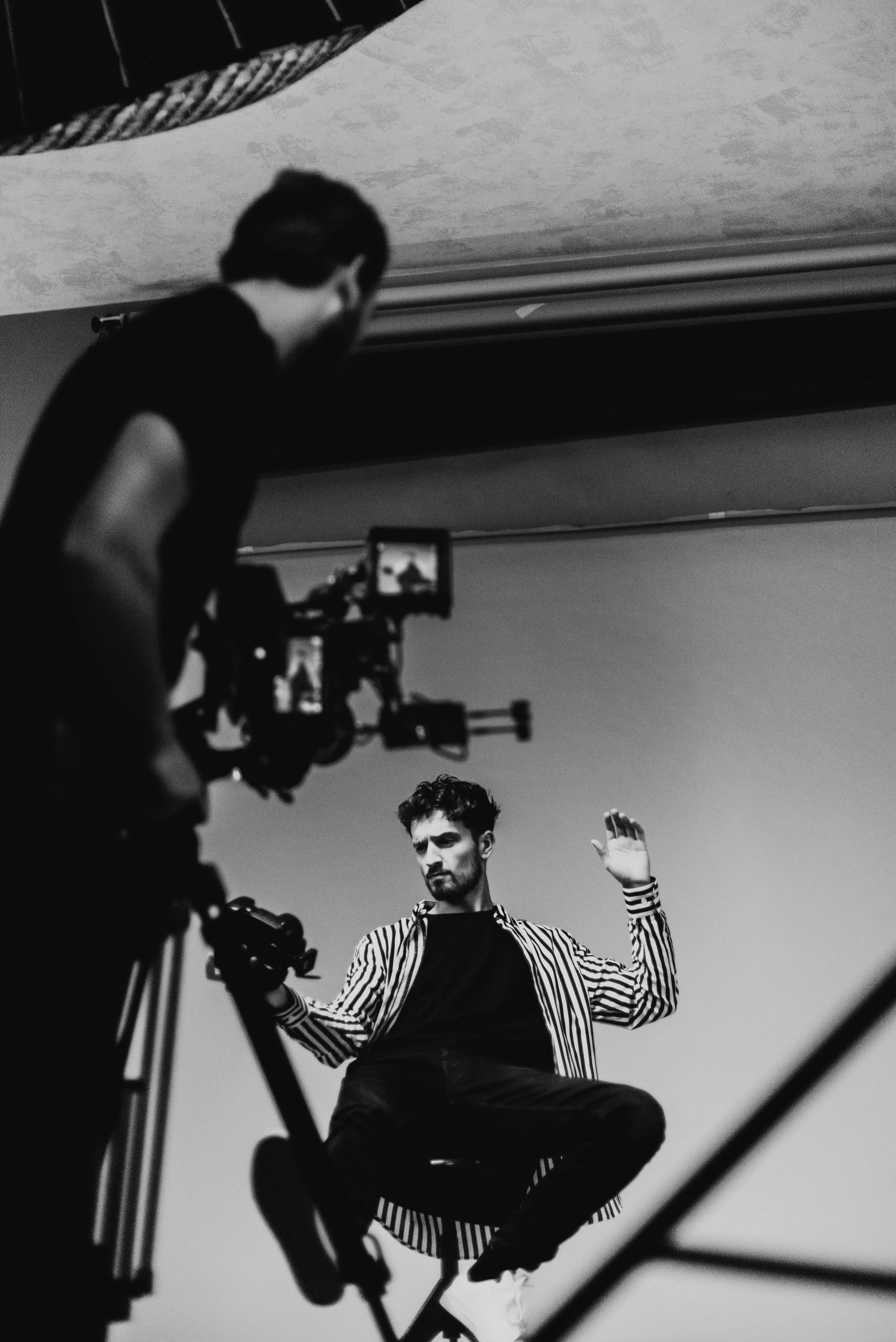 A man with a serious expression sitting on a stool with his left hand raised, being filmed by a person operating a camera, in a black-and-white photo.