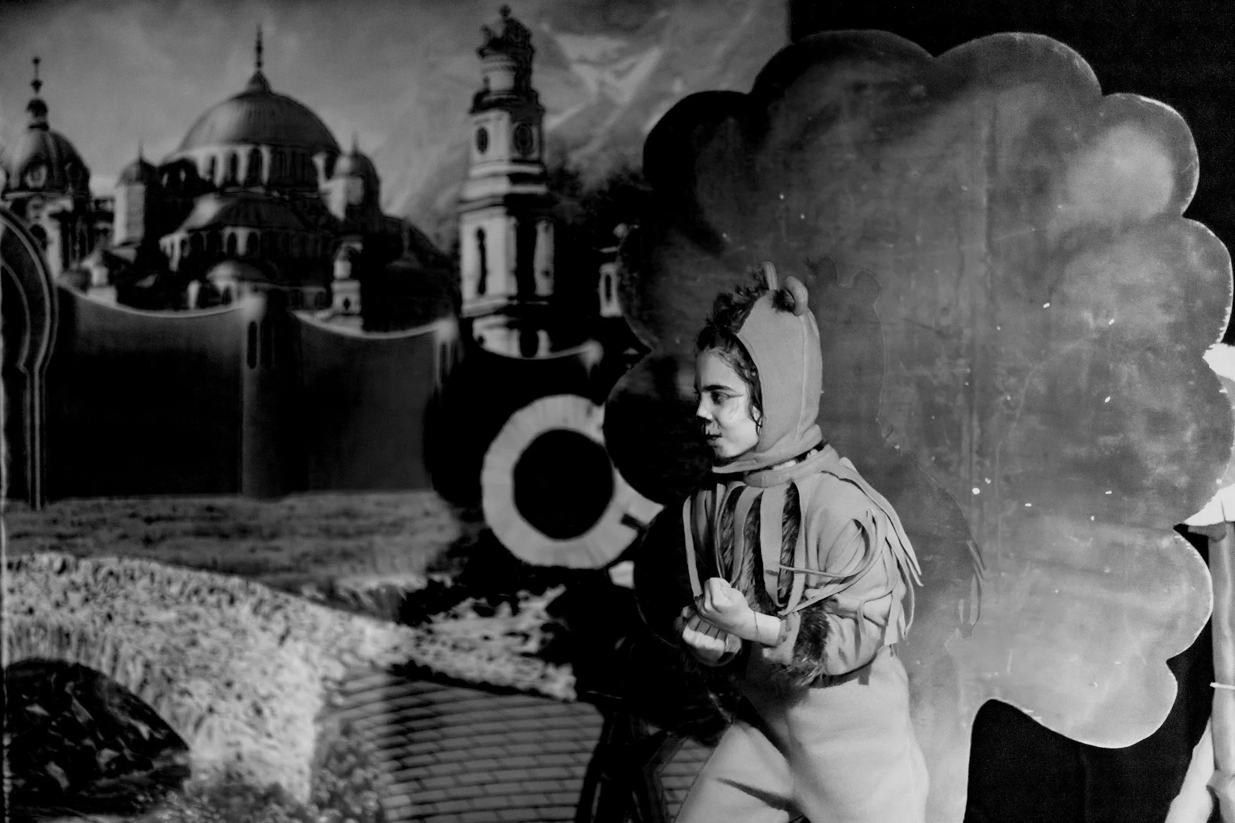 A black and white photo of a person in a costume, resembling a bear, standing in front of a painted backdrop of a castle and clouds.