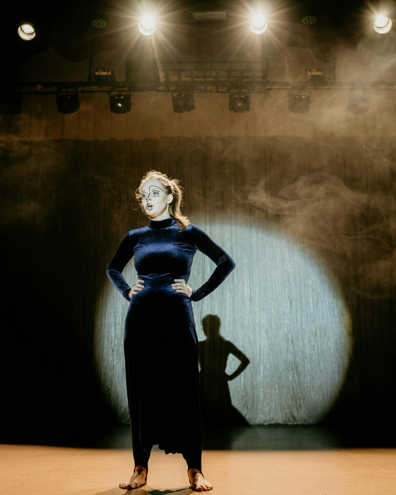 A woman on stage with dramatic lighting, dressed in a dark, velvet gown, standing with hands on hips, with makeup and glasses, with her shadow cast on the curtain behind her.
