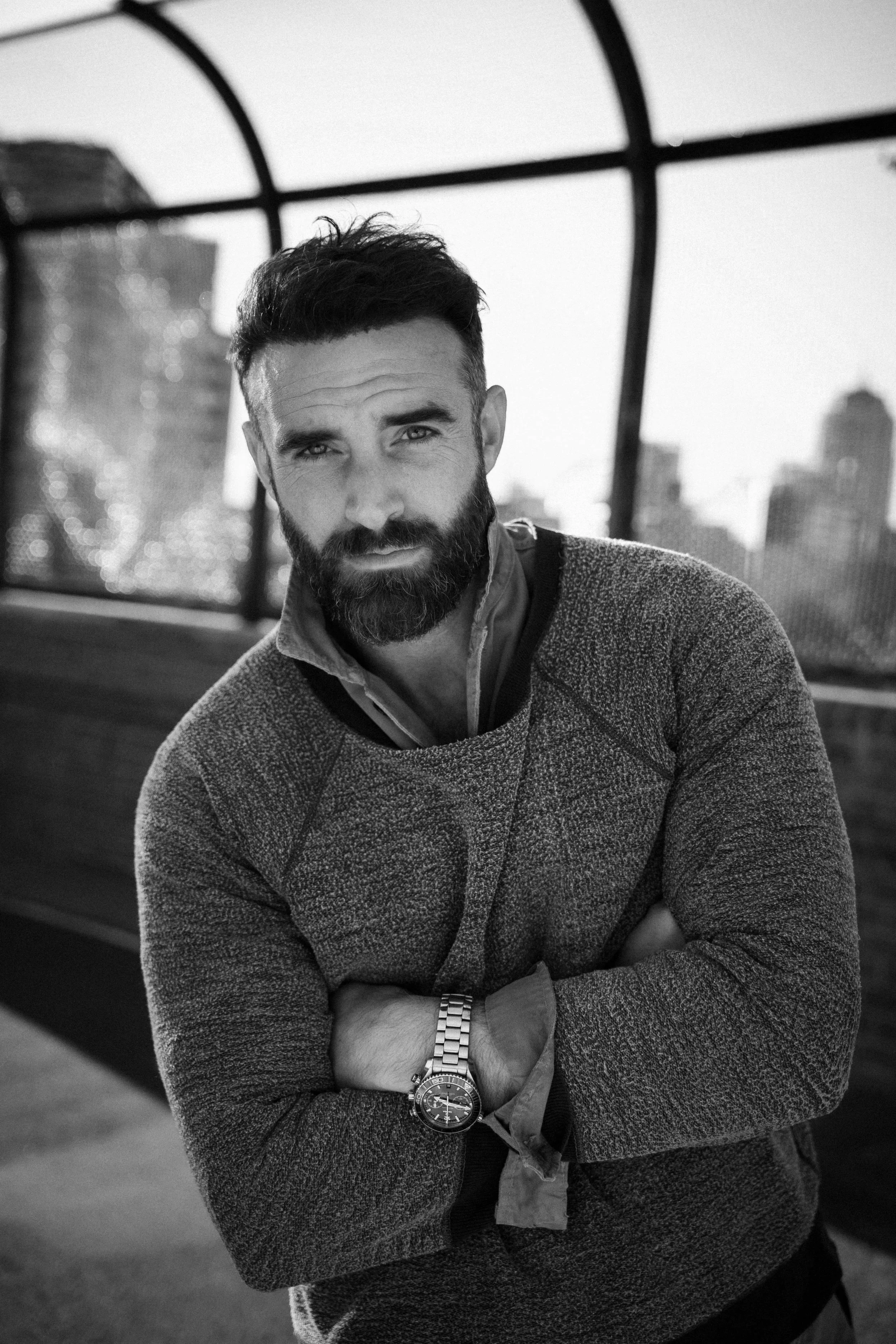 A black and white portrait of a man with a beard and short, styled hair. He is wearing a textured sweater, a collared shirt, and a watch, with his arms crossed. The background features a city skyline with tall buildings and a wire structure, suggesting an outdoor urban setting.