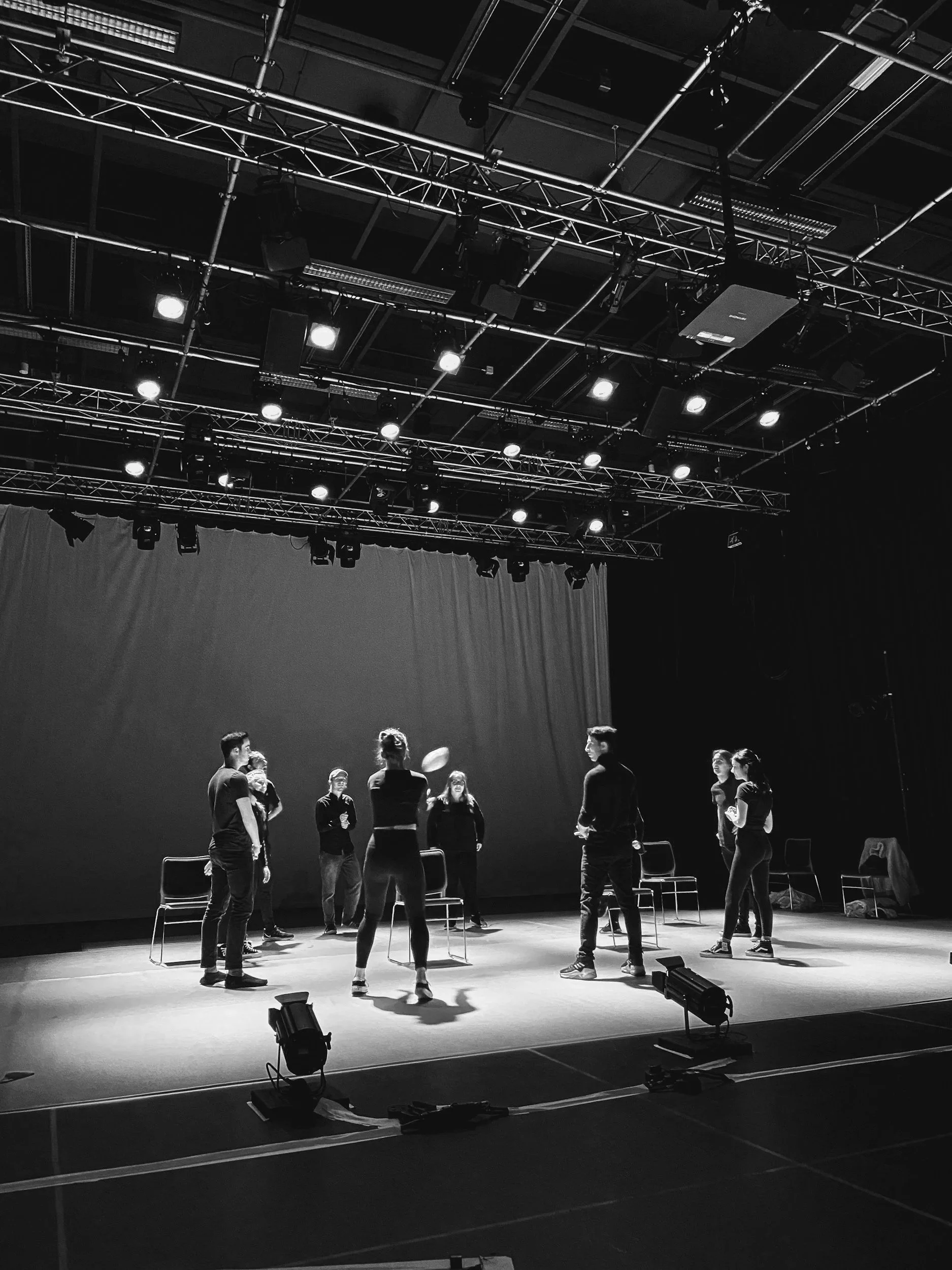 Black and white photo of people on stage rehearsing for a play or performance, with chairs and stage lights overhead.