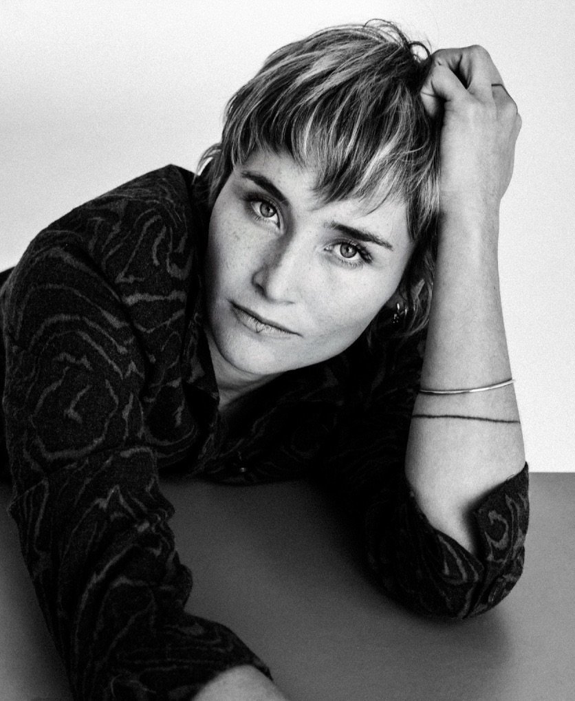 Black and white photo of a woman with short hair, resting her head on her right hand, looking directly at the camera.