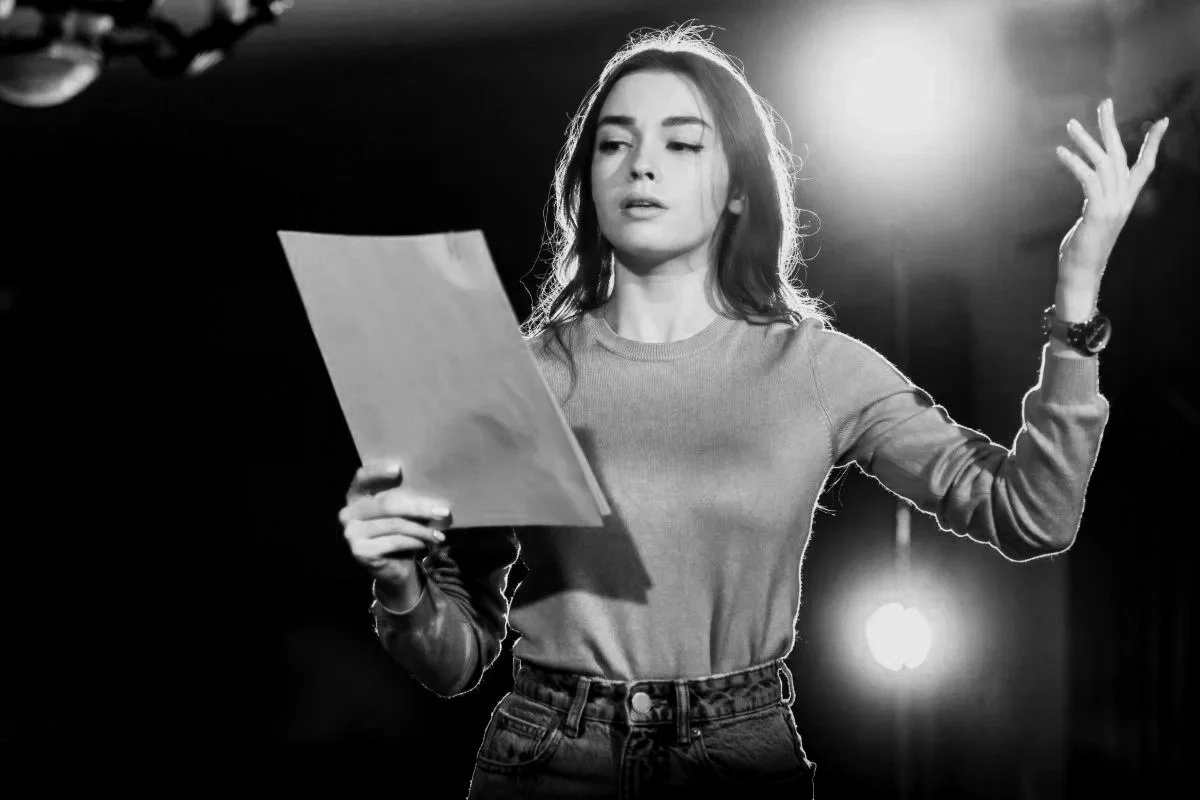 A woman with long hair reading a script or paper on a stage, gesturing with one hand, illuminated by stage lighting.