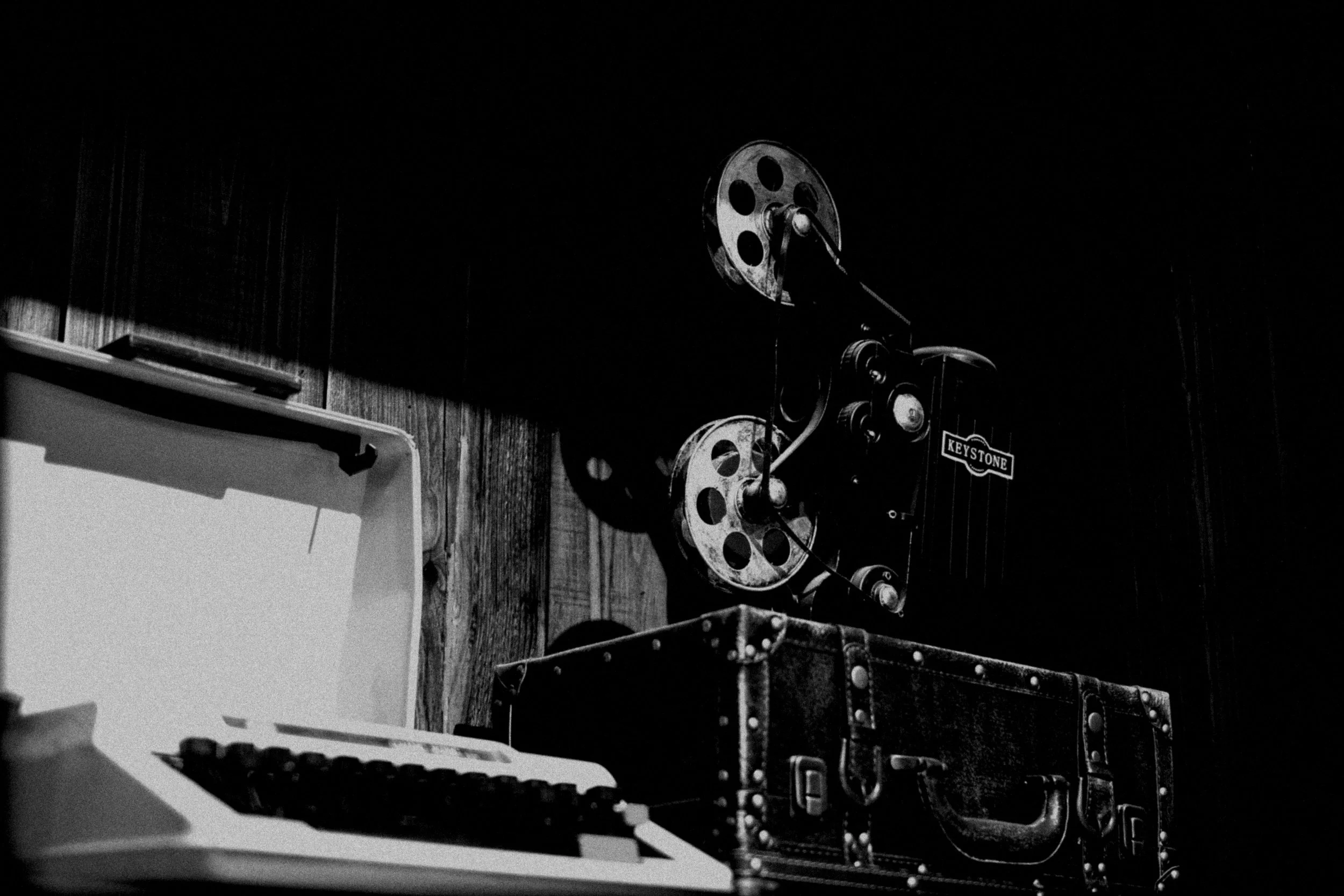 Vintage film projector with the brand name 'Keystone' on a wooden-paneled wall, with a typewriter and a small suitcase nearby in black and white.