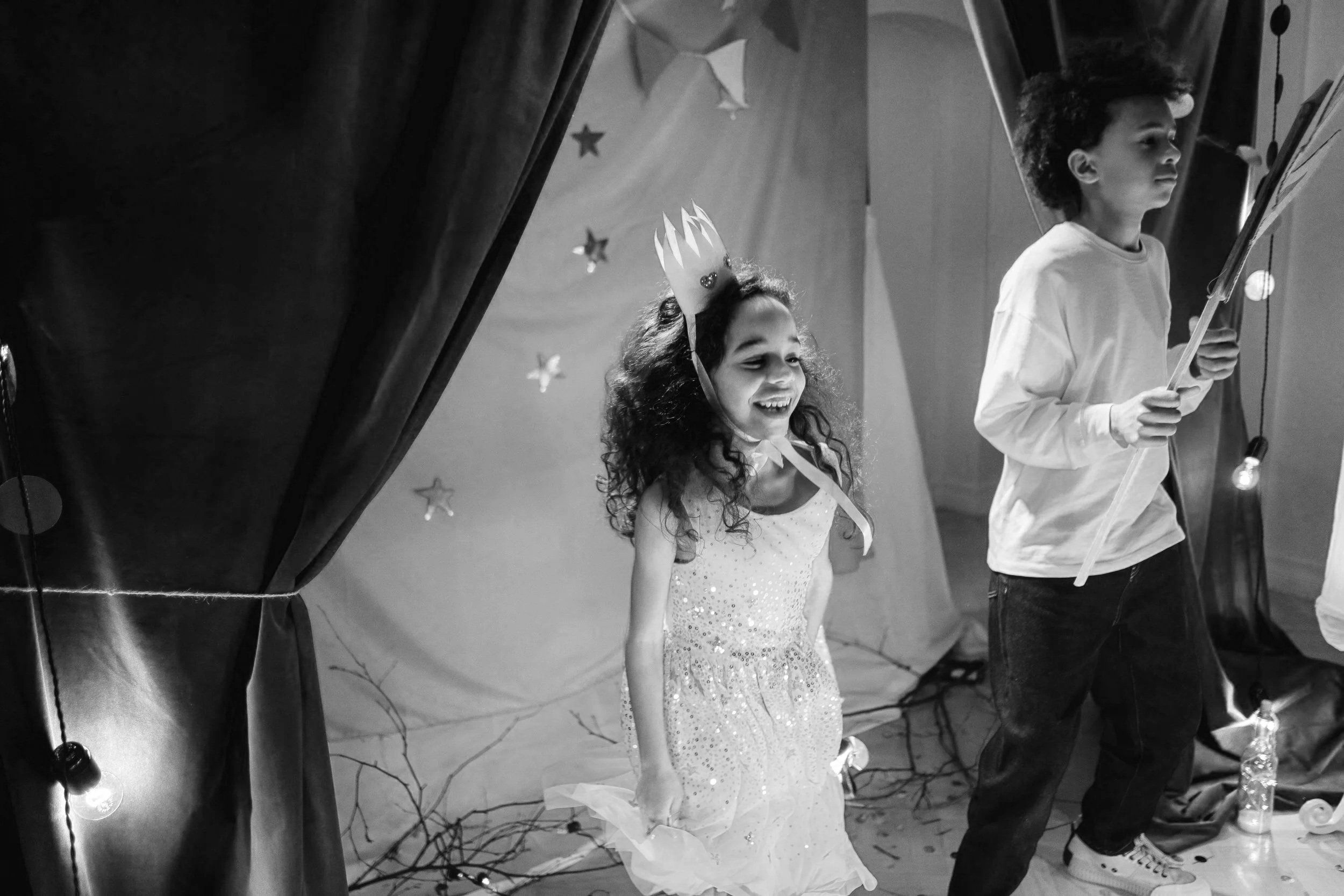 Two children performing on stage, a girl wearing a crown and a sparkly dress, smiling, and a boy holding a stick, possibly a prop, with curtains and stage decorations in the background.