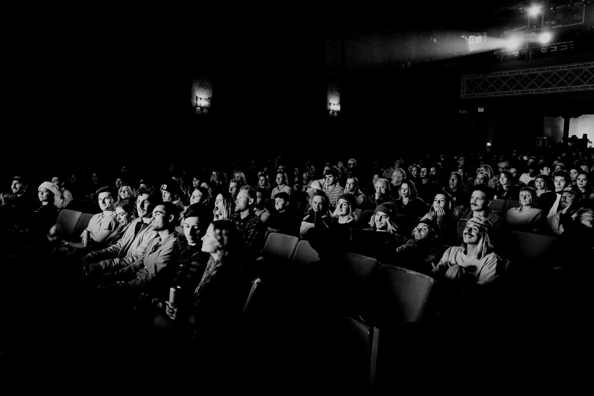 Black and white photo of a theater audience watching a show or movie, with some people smiling or focused on the stage.