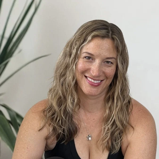 A woman with long, wavy blonde hair smiling at the camera, wearing a black top and a star-shaped necklace, sitting indoors near a plant.