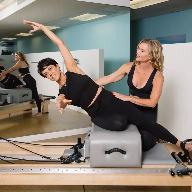 Woman performing Pilates exercise on a reformer machine while a trainer assists in a fitness studio with a mirrored wall.