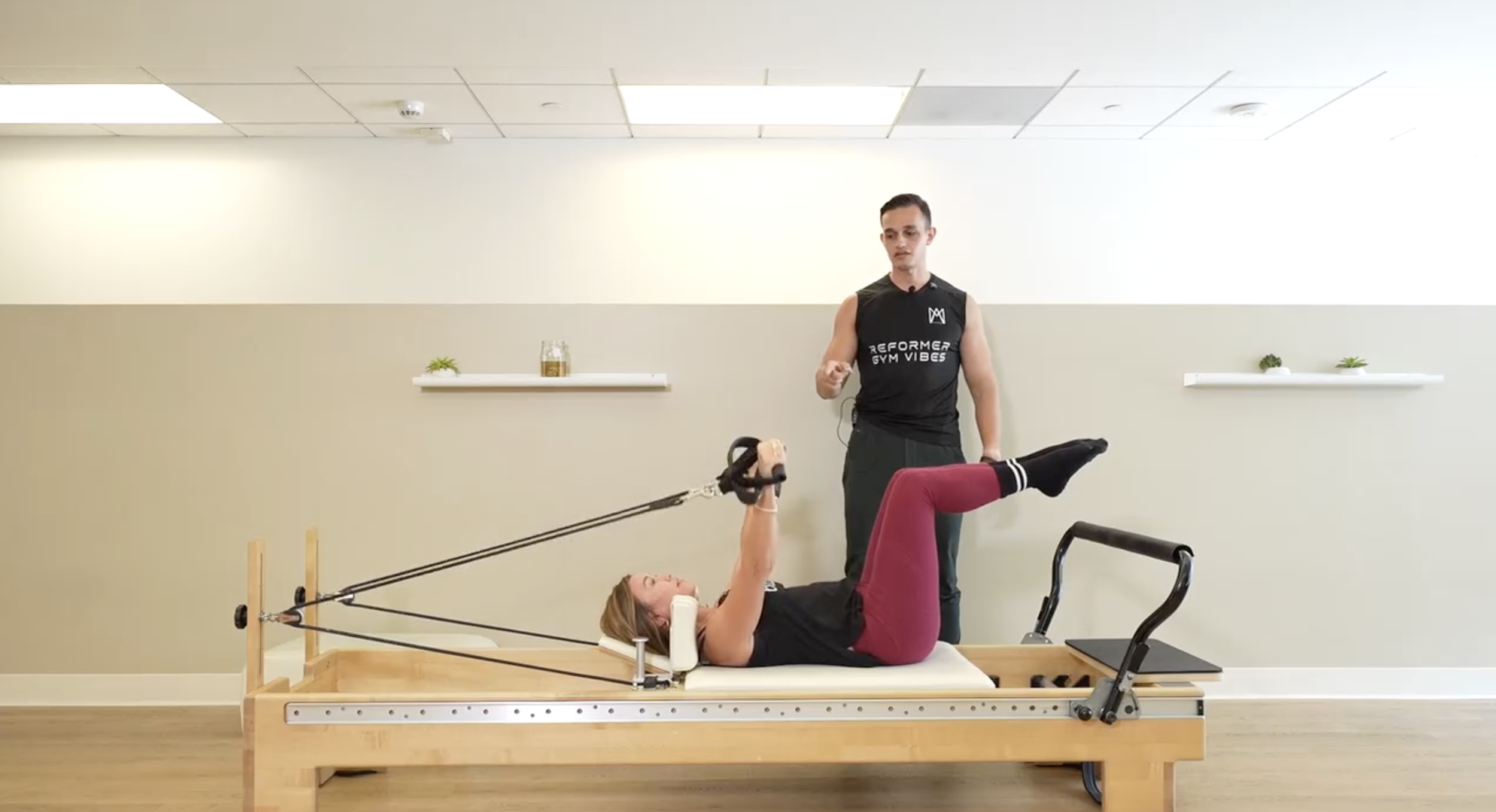 A woman lying on a Pilates reformer machine performing an exercise with resistance band, supervised by a male instructor, in a fitness studio.