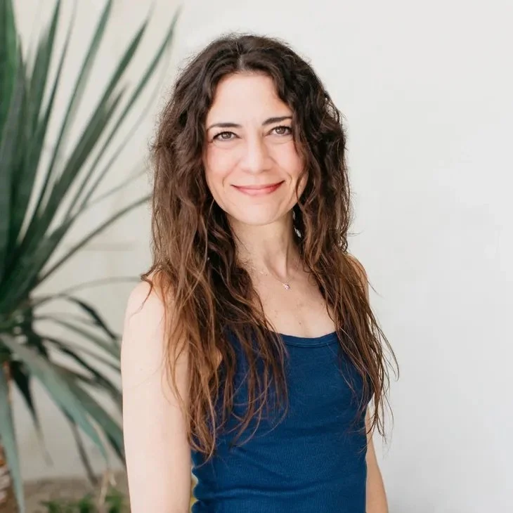 Smiling woman with long, wavy brown hair wearing a blue tank top, standing near a large green plant with a white background.
