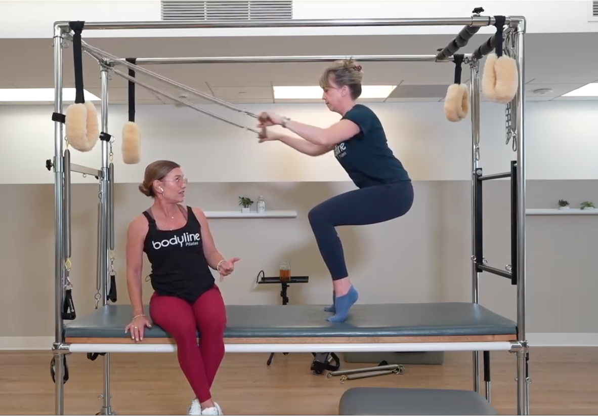 A woman with glasses, wearing a black tank top with 'bodyline' written on it and red leggings, is sitting on a platform. Another woman in a dark shirt and navy leggings is performing an exercise on a Pilates reformer machine, jumping with knees bent and arms extended forward, while wearing socks. The scene is in a fitness studio with minimal decor.