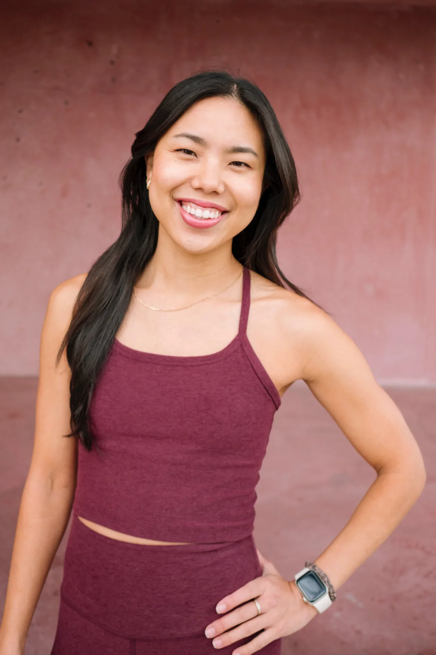 A young woman with long dark hair, smiling, wearing a sleeveless maroon athletic outfit and an Apple Watch, standing in front of a reddish-brown wall.