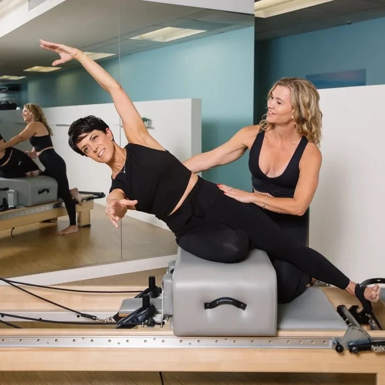Two women exercising with a Pilates reformer machine in a fitness studio. One woman is lying sideways on the machine, stretching her arm upward, while the other woman, nearby, supports her and smiles.