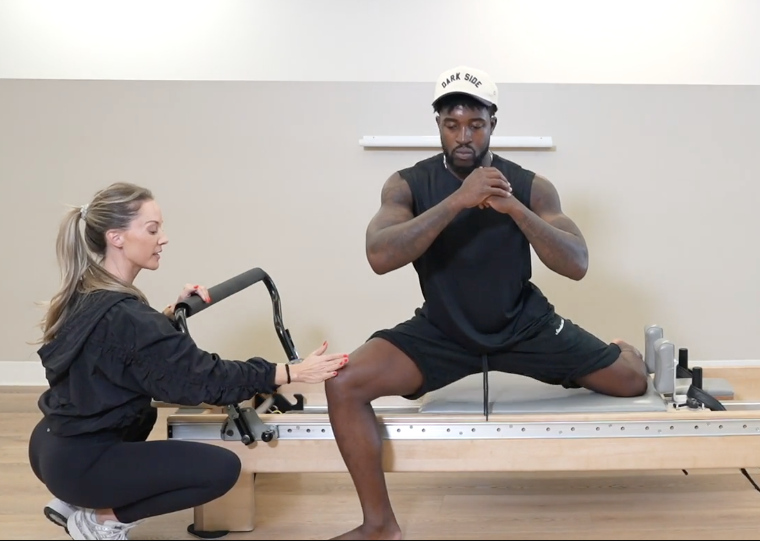 A person with tattoos practicing a fitness move with a trainer assisting, another woman kneeling and holding the person's leg, in a gym or fitness studio.