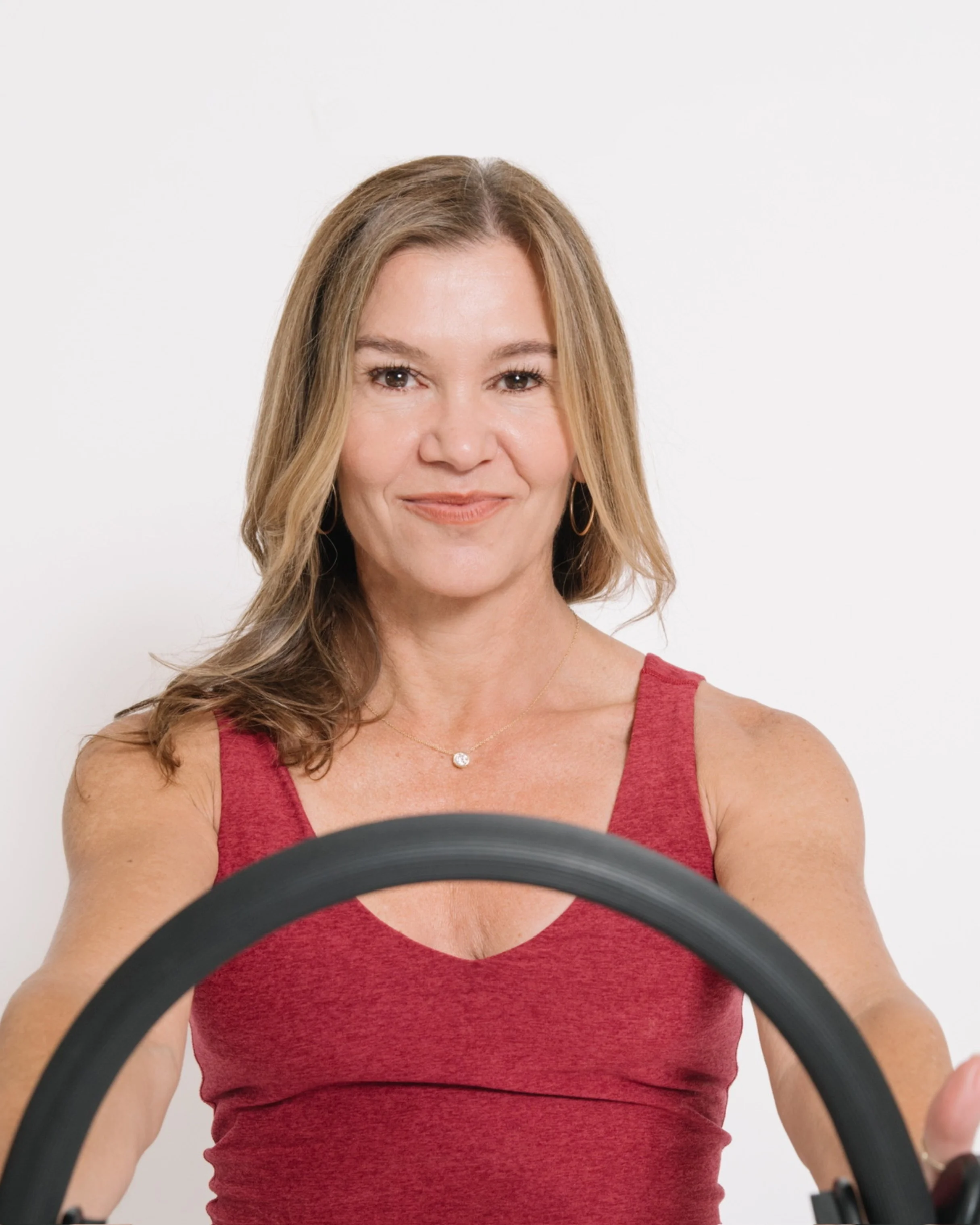Woman with light brown hair and hoop earrings smiling, wearing a sleeveless red top, holding a steering wheel, against a plain white background.