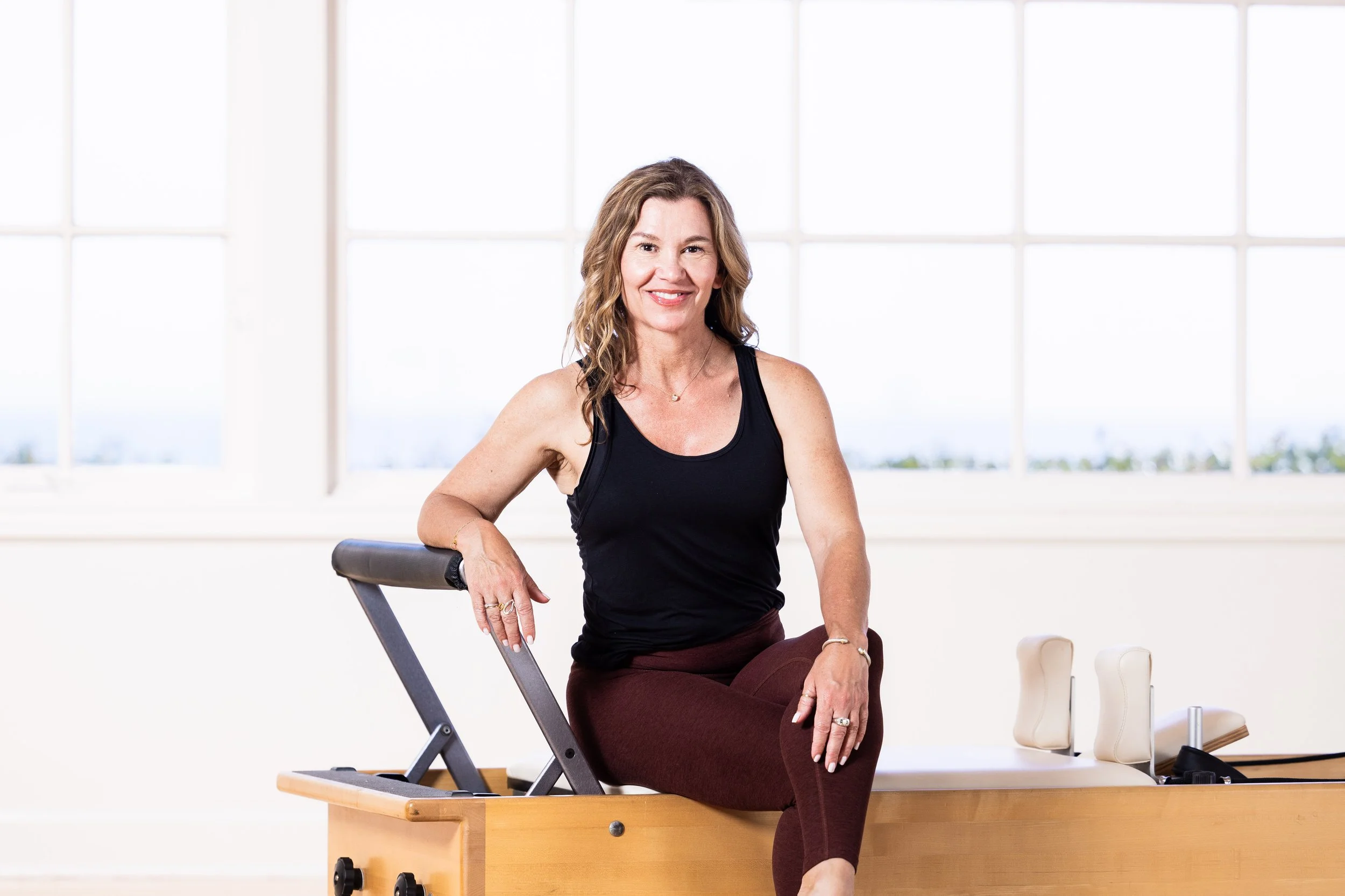 A woman sitting on a Pilates reformer machine, smiling, in a bright room with large windows in the background.