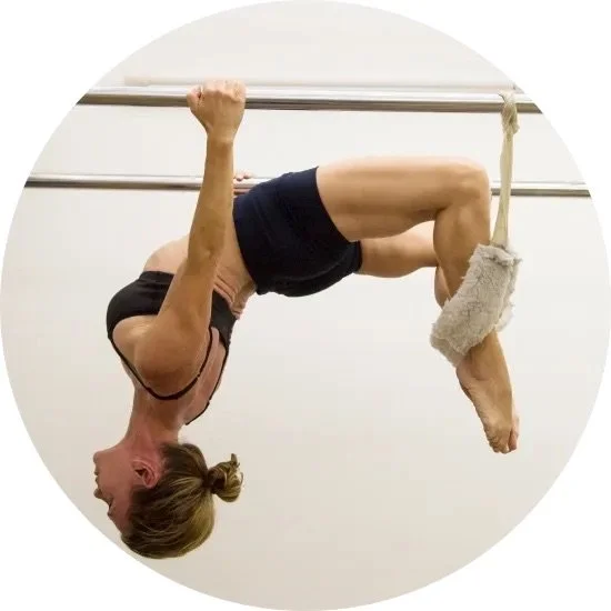 An elderly woman performing a neckstand or yoga pose while hanging upside down from ballet bars, with her feet wrapped in cloth and supporting her body.
