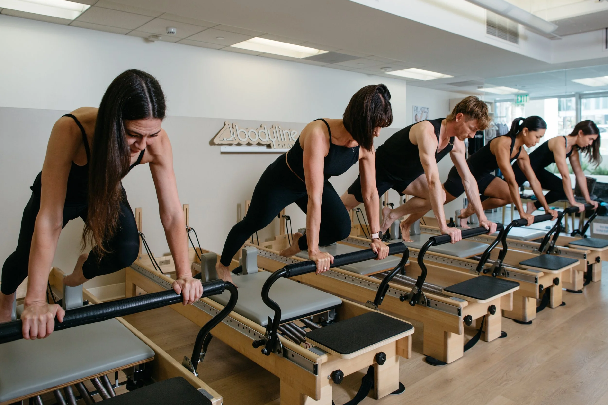A group of people dressed in black workout clothes are doing pilates on reformer machines in a fitness studio. They are in a plank position, leaning forward with their hands on the reformers. The studio has a sign on the wall that reads 'Abodyline Pilates' and large windows letting in natural light.