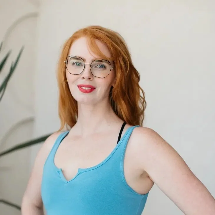 A young woman with red hair, glasses, and red lipstick wearing a blue tank top, standing indoors against a light-colored wall with a plant in the background.
