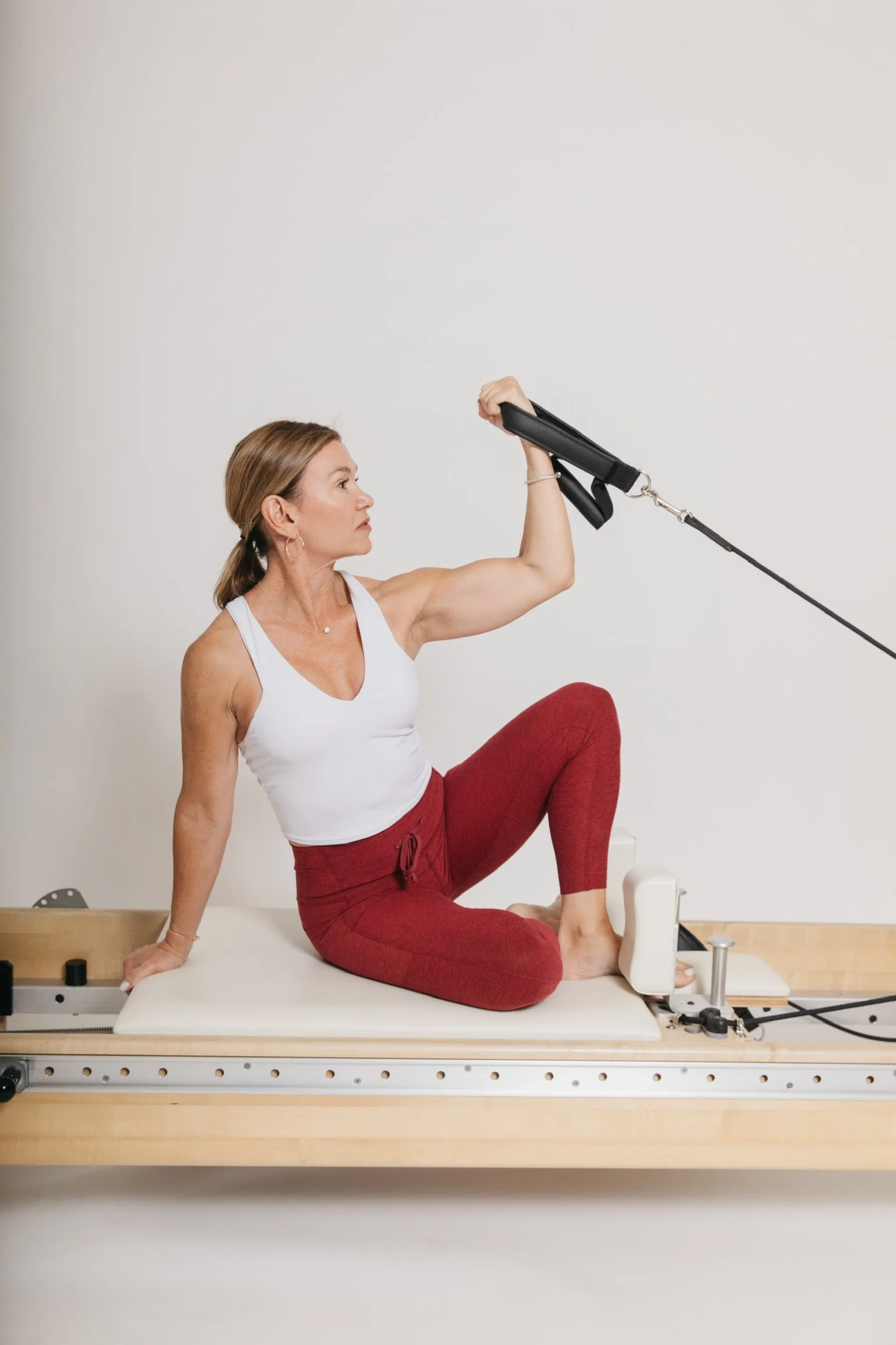 Woman exercising on Pilates reformer machine, sitting with one arm raised, in a white tank top and red leggings, in a gym or fitness studio.
