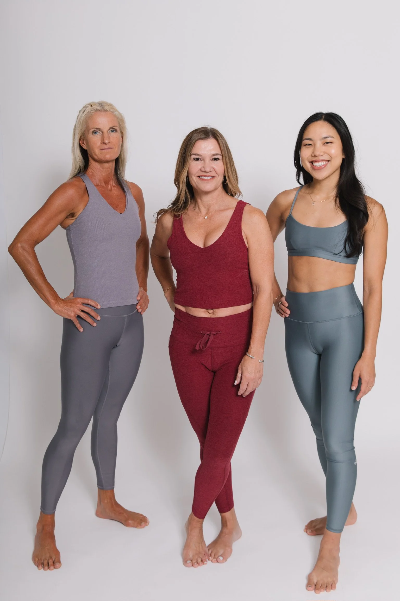 Three women standing barefoot against a white background, dressed in activewear. The woman on the left has long gray hair and wears a purple tank top with gray leggings. The woman in the middle has shoulder-length blonde hair and wears a red sports bra with matching red pants. The woman on the right has long black hair and wears a blue sports bra with matching blue leggings. All are smiling.