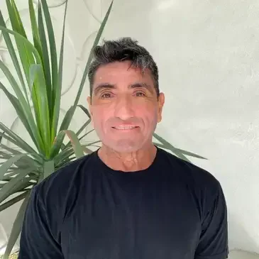 Man smiling, standing inside next to a plant wearing a black tee shirt