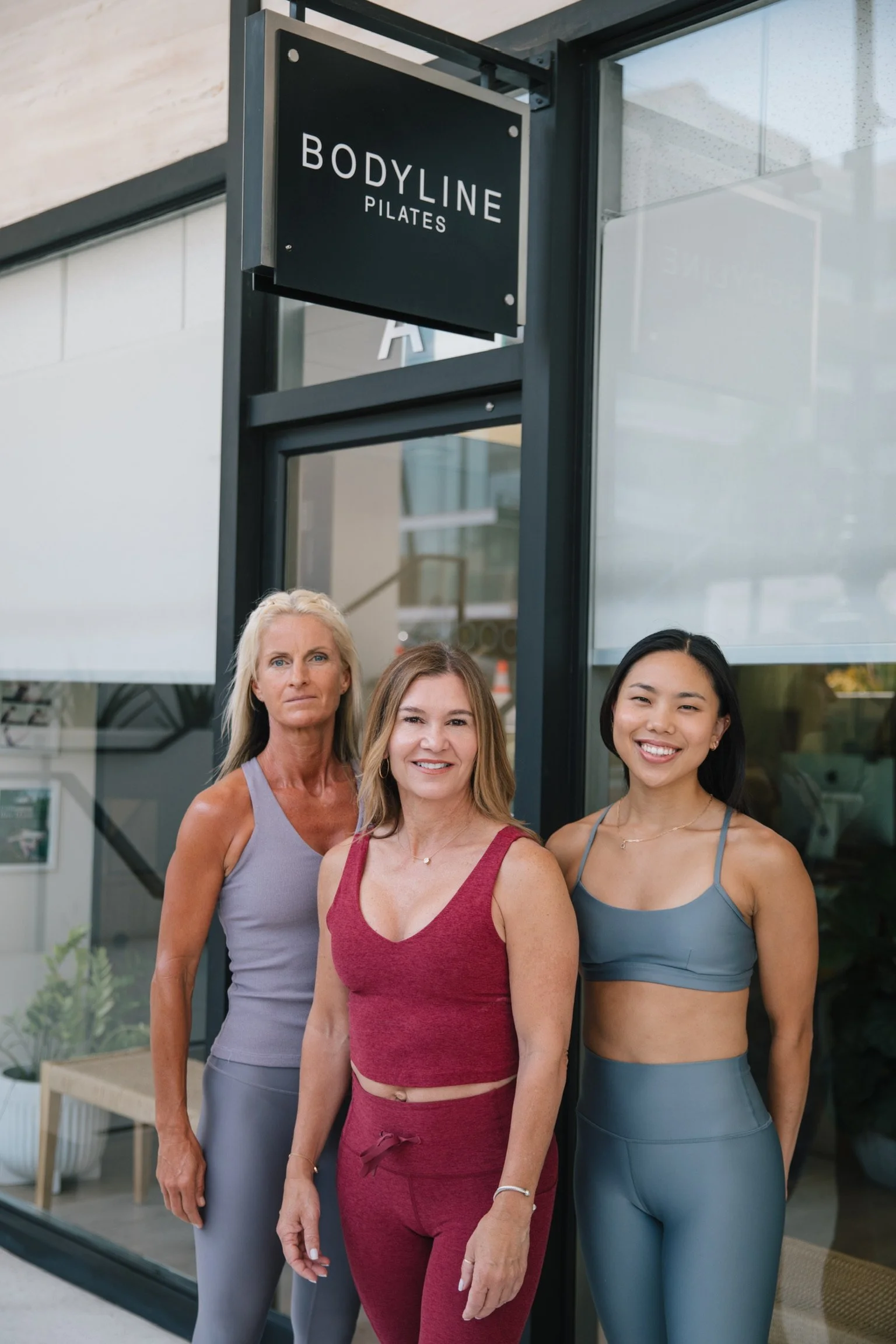 Three women standing outside of a pilates studio called 'Bodyline Pilates.' They are dressed in athletic wear and smiling.