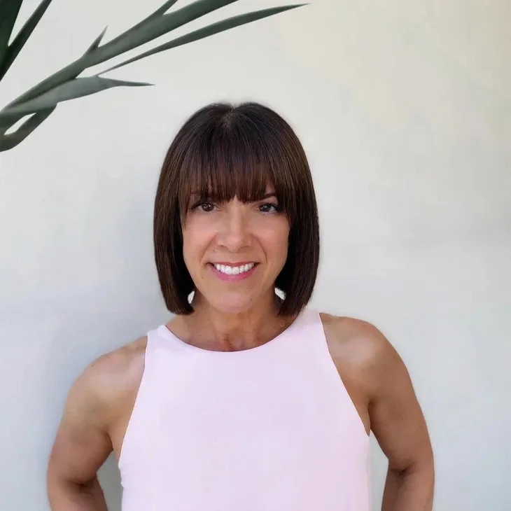 Woman with shoulder-length dark brown hair and bangs, smiling, wearing a sleeveless light pink top, standing in front of a light-colored wall with a green plant in the upper left corner.