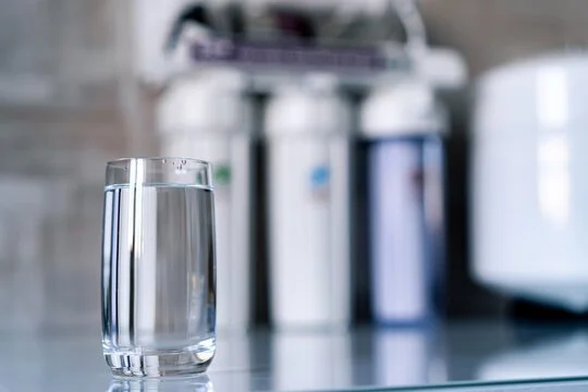 A clear glass of water on a reflective surface in a kitchen with appliances in the background.