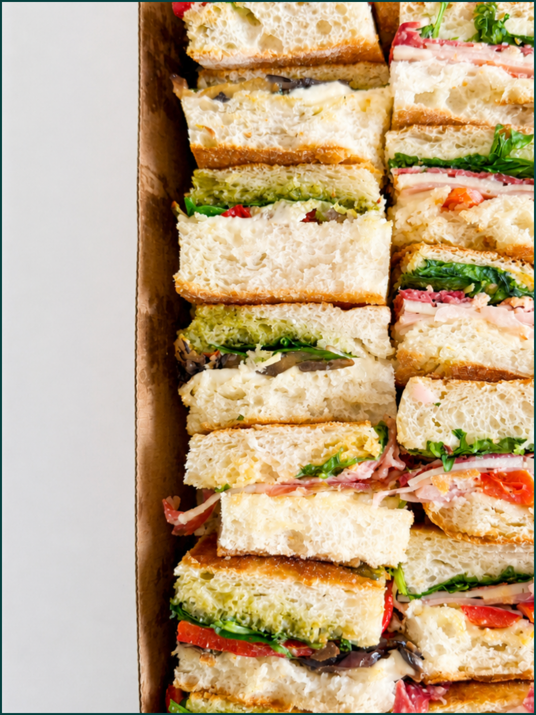 Assorted focaccia panini with various fillings such as lettuce, tomatoes, meats, cheese, and spreads, arranged in a cardboard box for catering