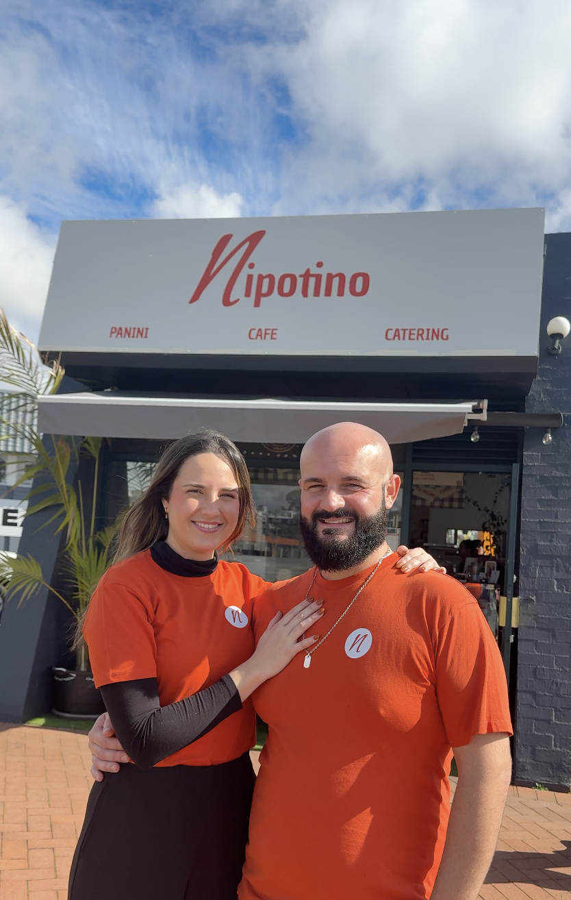 Marta and Antonio are standing outside a restaurant named Nipotino, with the woman touching the man's chest. The restaurant offers panini, cafe, and catering, as indicated on the sign above them.