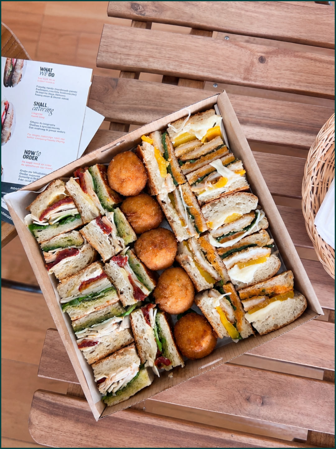 A catering box filled with assorted panini and fried snack balls on a wooden table.