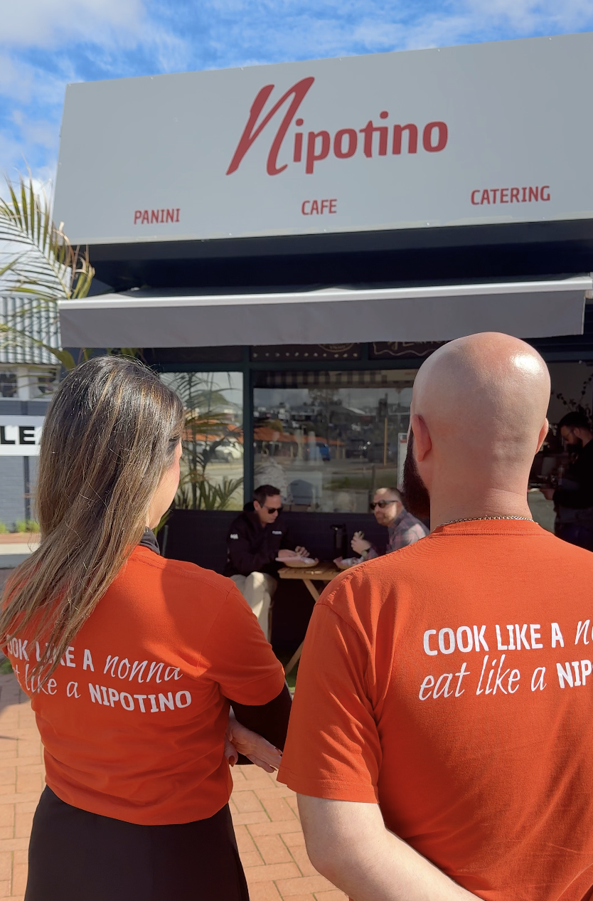 Marta and Antonio are wearing matching orange shirts with the phrase "COOK LIKE A NONNA EAT LIKE A NIPOTINO" on the back, standing outside of a restaurant named Nipotino, with a few people sitting at a table inside.
