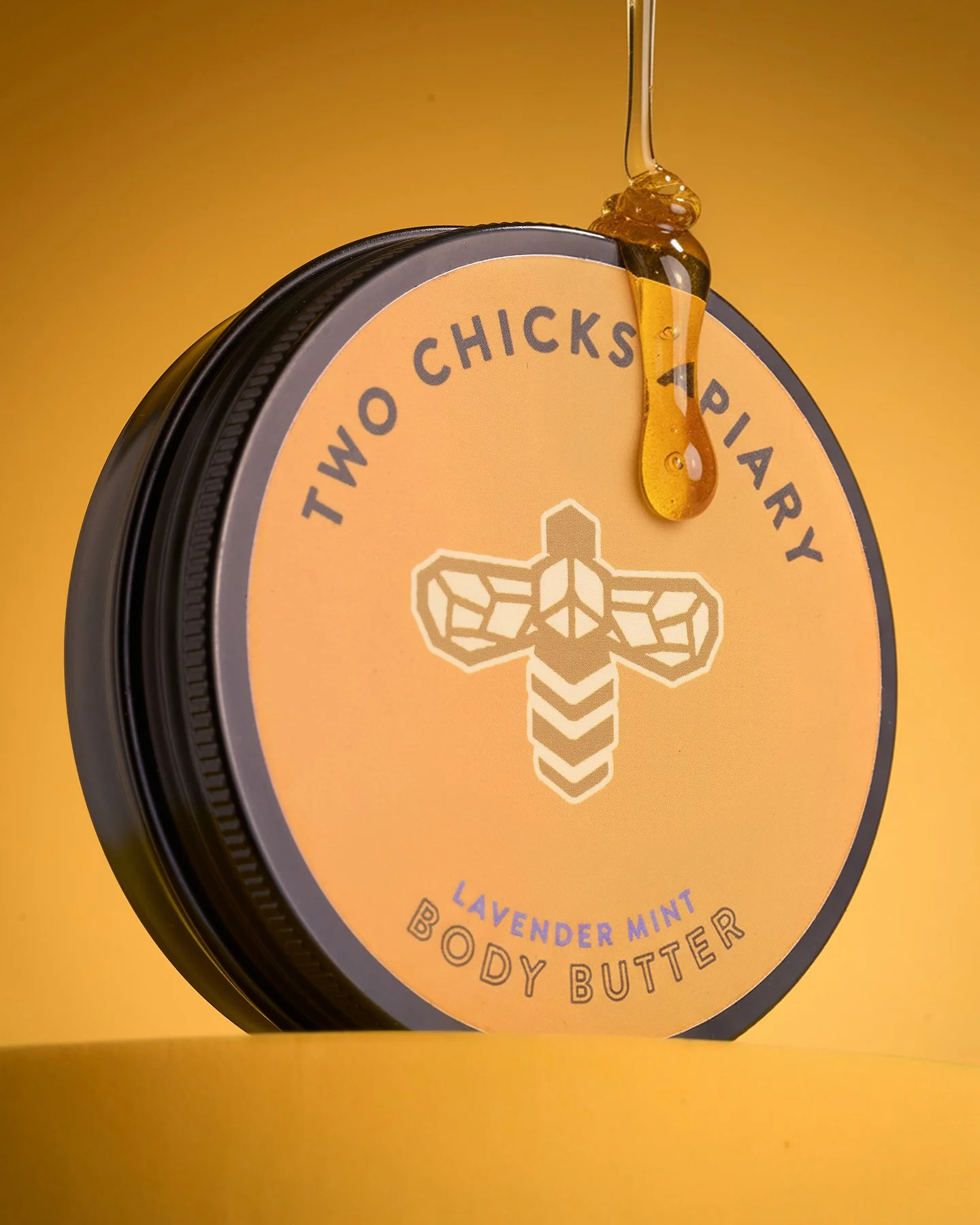 Close-up of a container of Two Chicks Pharmacy Lavender Mint Body Butter with honey or oil dripping from a spoon above it, against a matching yellow background.