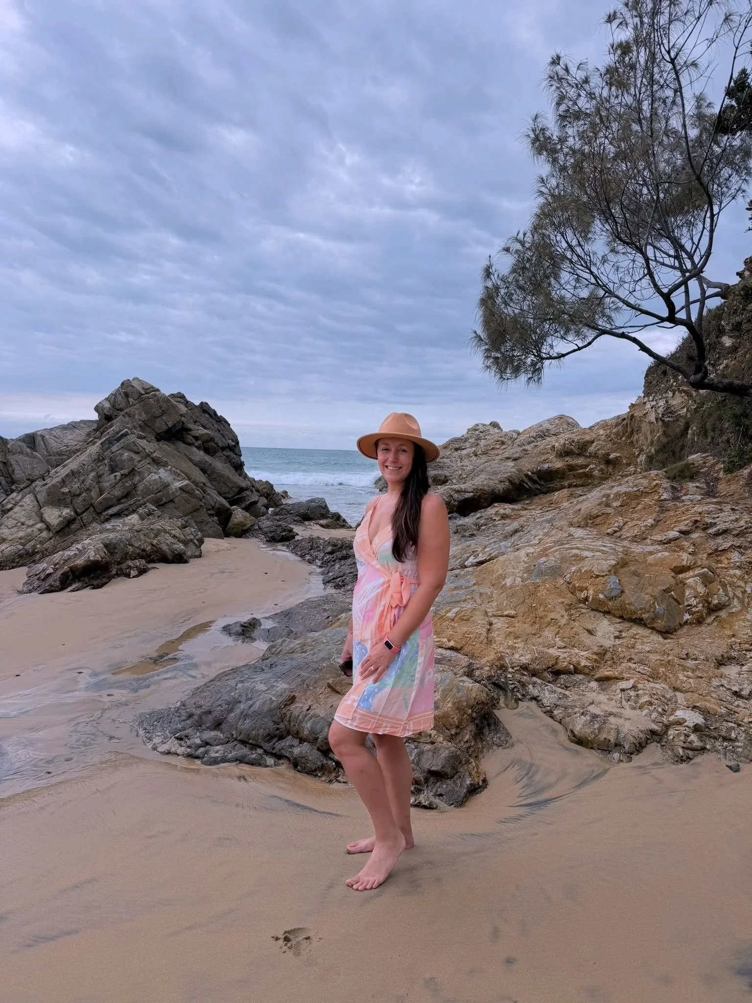 A woman in a colorful dress and wide-brimmed hat standing barefoot on a sandy beach with rocks, trees, and ocean waves under a cloudy sky.